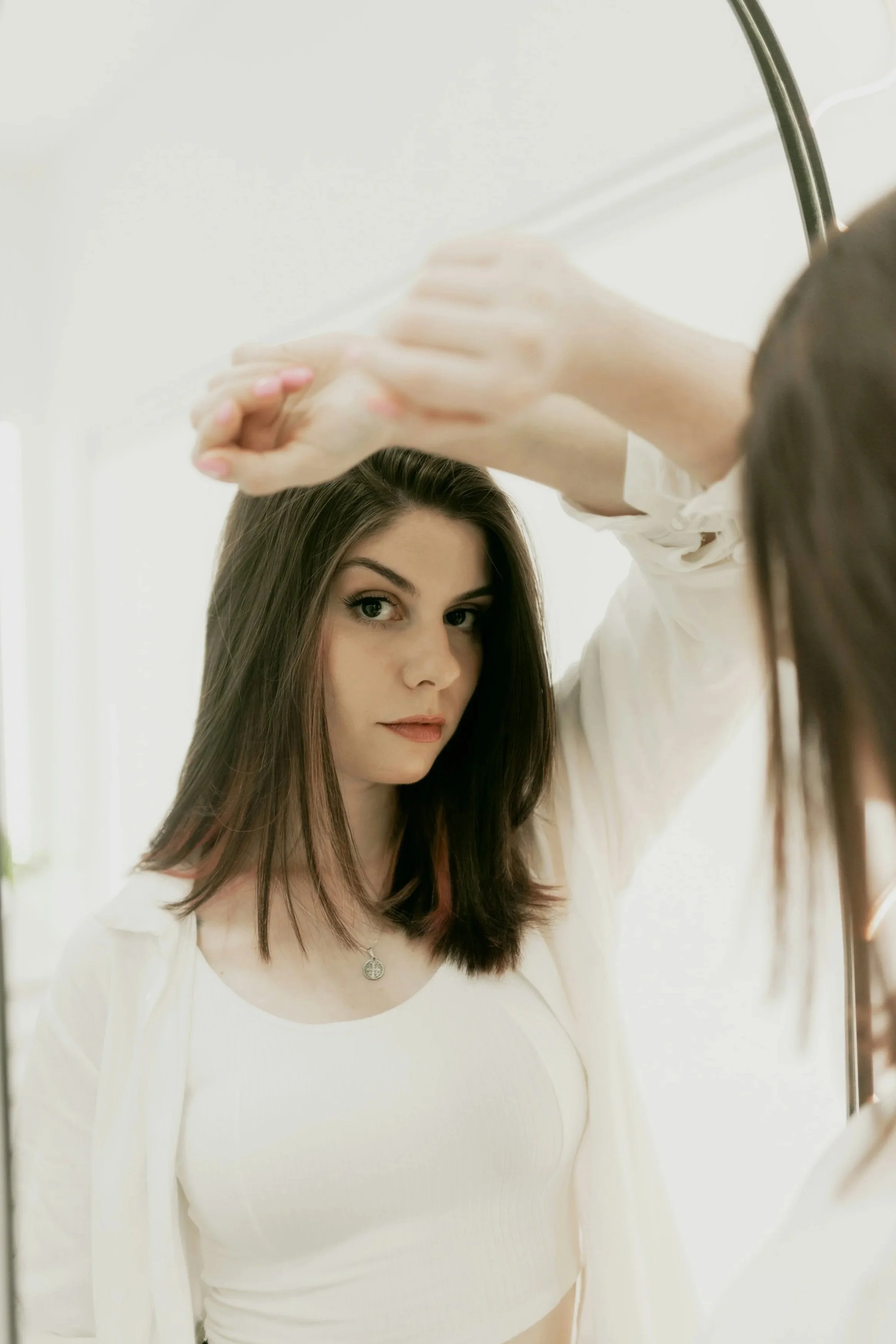 A young woman with dark brown hair and fair skin looking at herself in a mirror, wearing a white top, with her hand raised to her forehead, in a well-lit room.