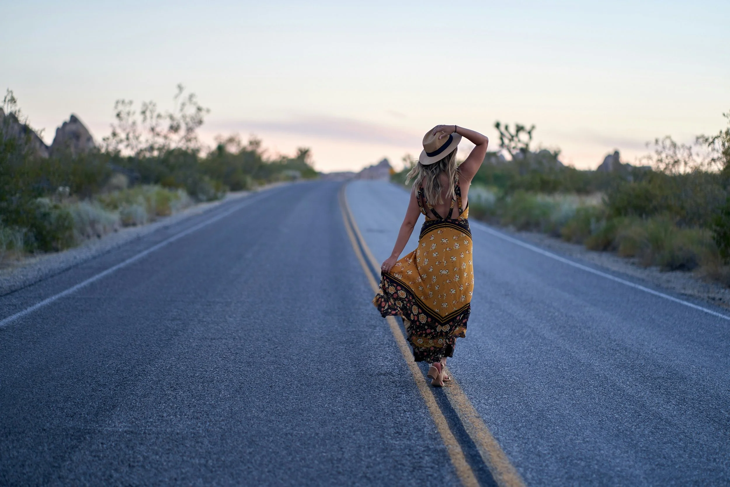 A woman in a bohemian dress and a hat walking down the center of a deserted road in a desert landscape during sunset.