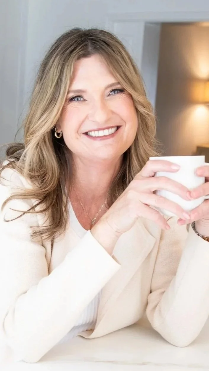 Smiling woman with wavy light brown hair and blue eyes holding a white mug, wearing a cream-colored blazer and jewelry, in a cozy indoor setting.