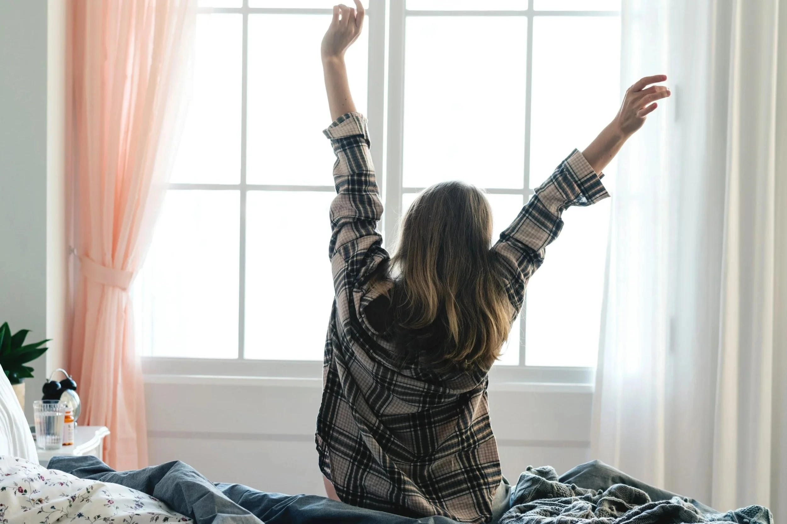 A woman sitting on a bed stretching with arms raised, facing a large window with white panes and pink curtains in a bright room.