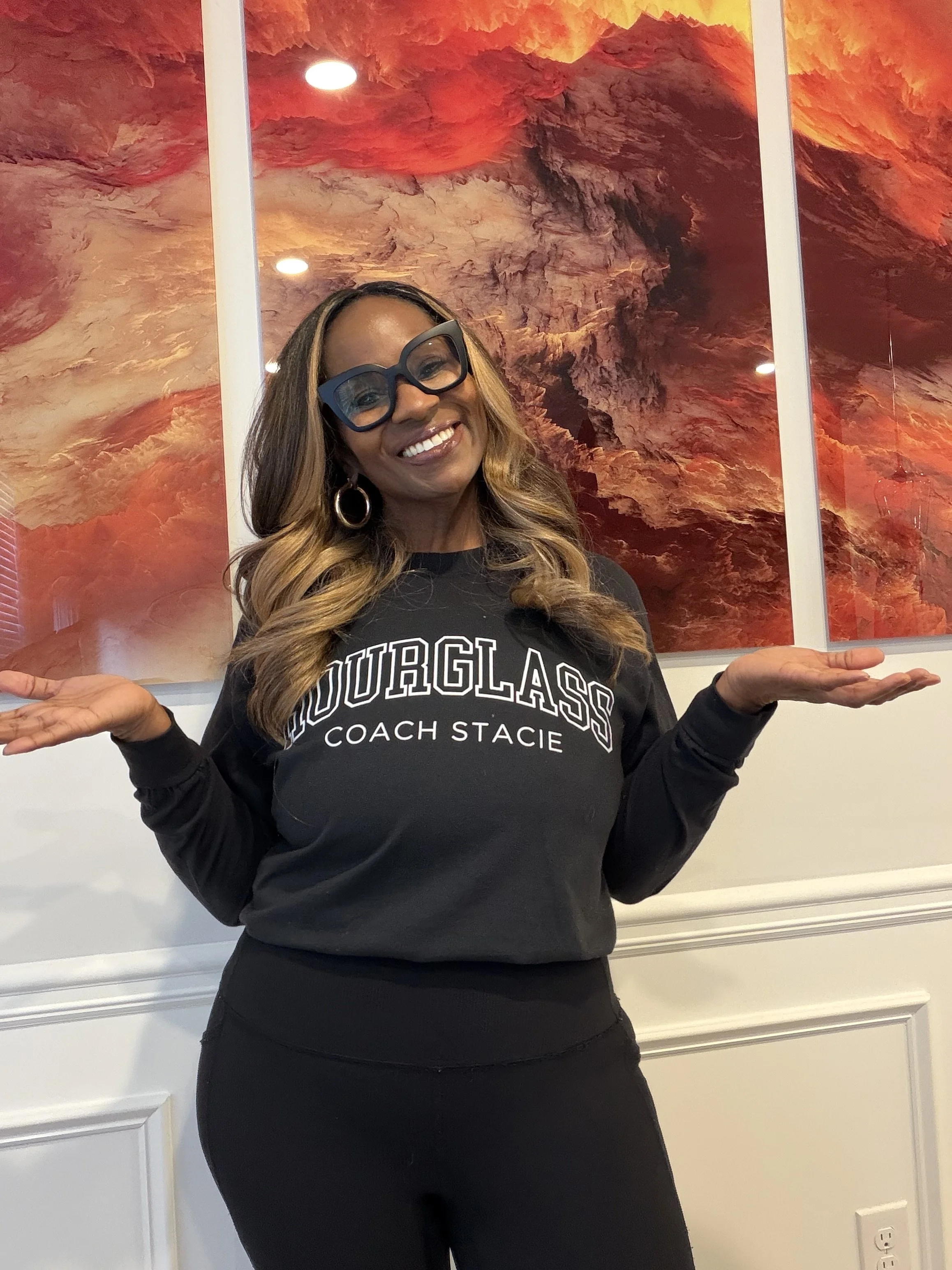 A woman with long wavy hair, wearing large blue glasses, hoop earrings, and a black sweatshirt that says 'YOURGLASS COACH STACIE' standing in front of an abstract red-orange artwork, smiling and gesturing with her hands.
