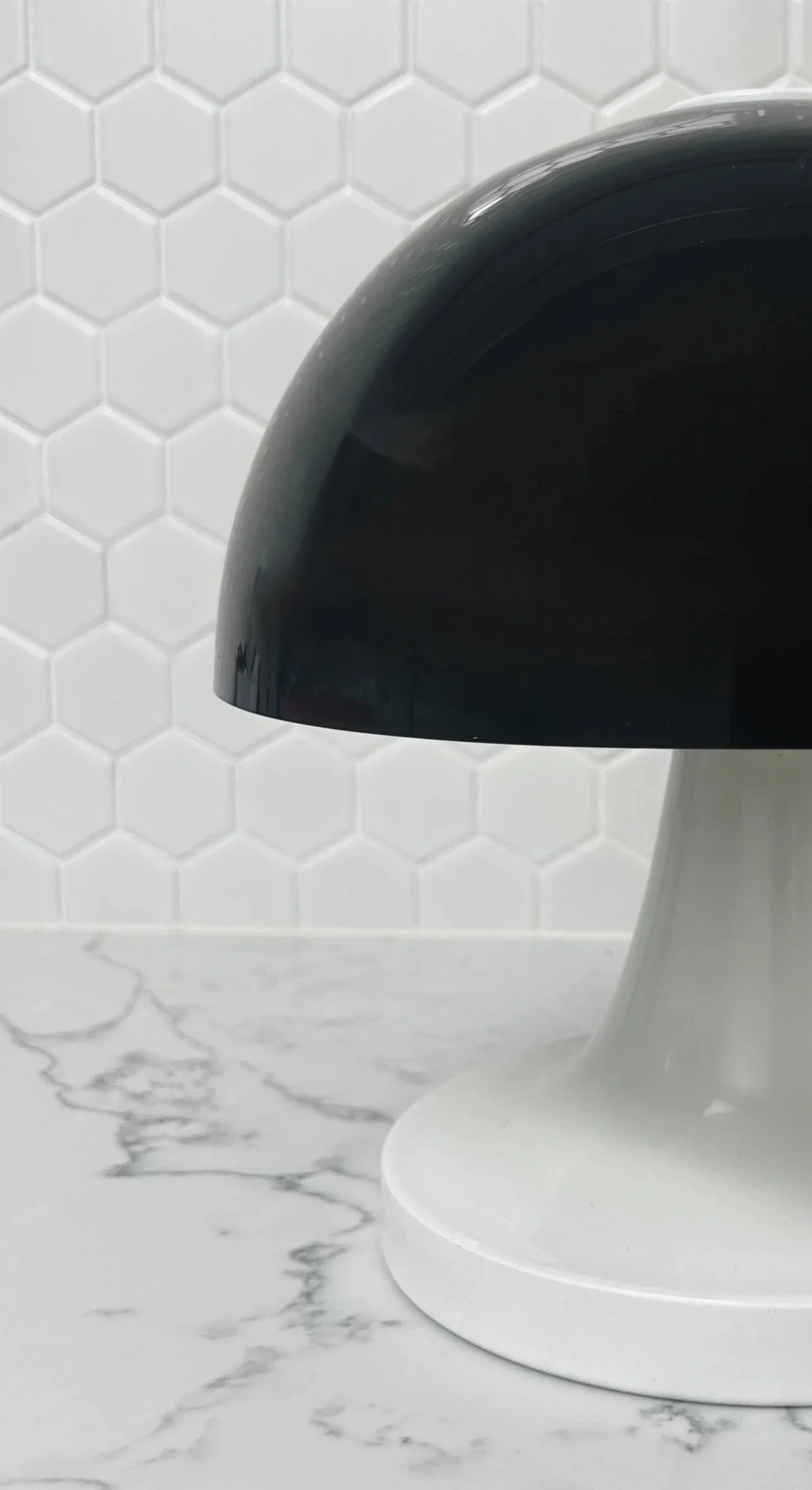 a mushroom table lamp with black acrylic dome shade sitting atop of a white enamel fluted base on a Mabel surface against a white hexagon tiled wall