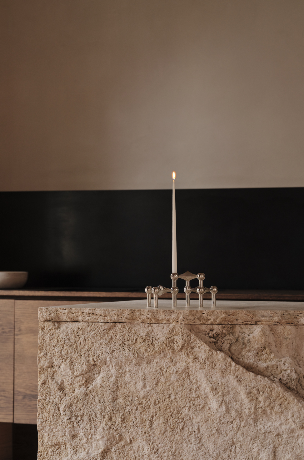 A lit candle on a selection of STOFF Nagel metal candle holders with spherical accents on a stone countertop in a minimalist interior.