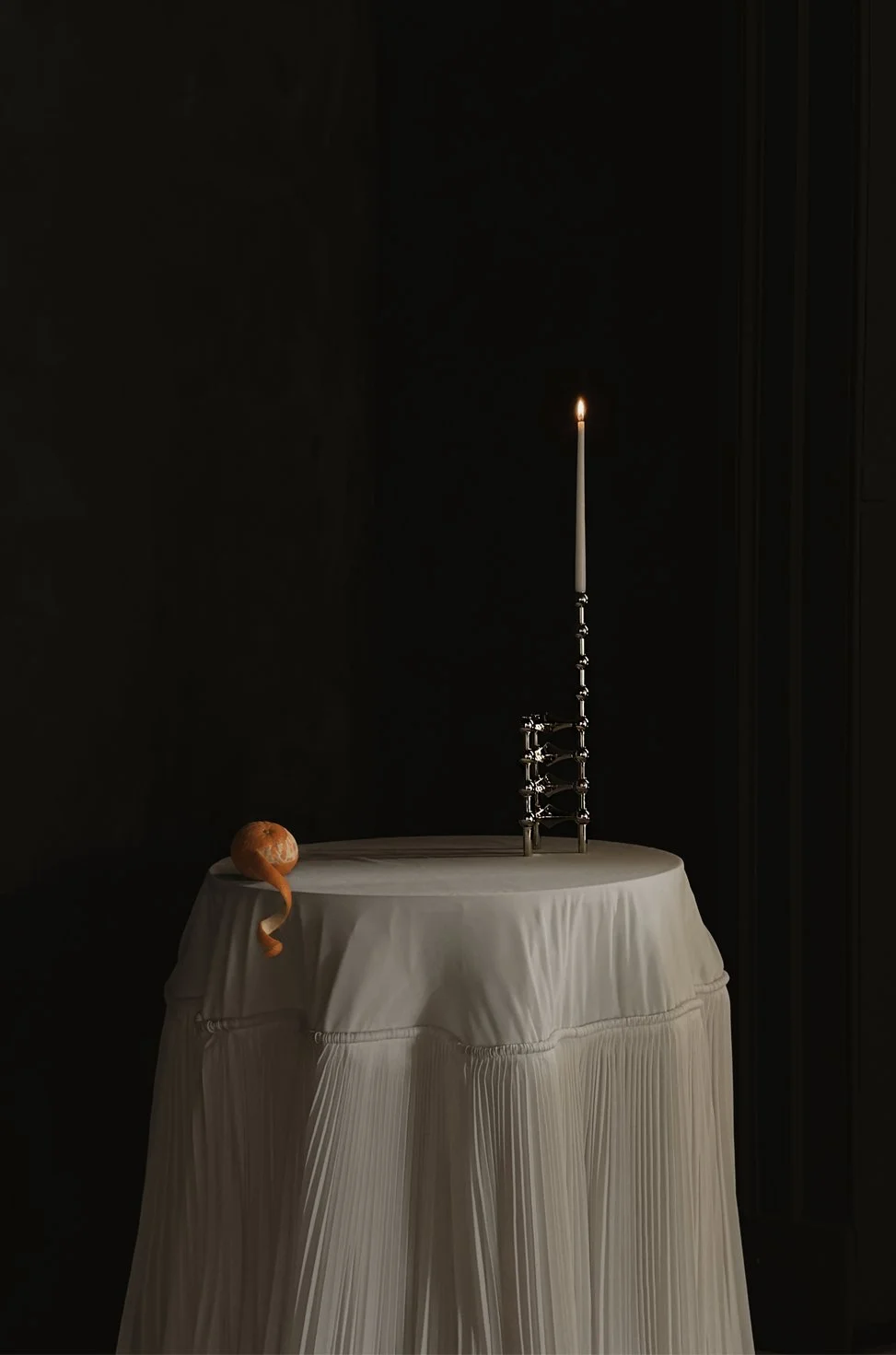 A single lit candle on a STOFF Nagel chrome candle holder, an orange with a peel partially removing itself, on a white tablecloth-covered round table against a dark background.