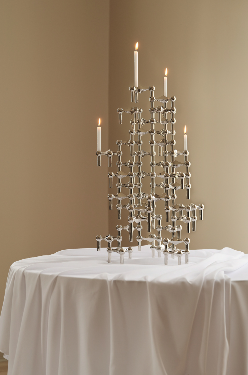 A collection of STOFF Nagel candle holders stacked on top of each other, with four lit candles on a table with white tablecloth.
