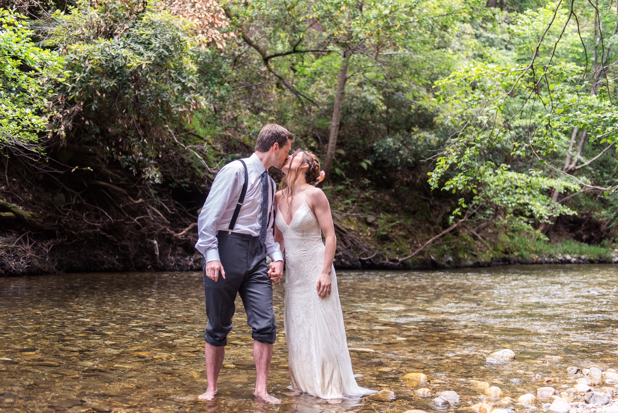 Couple kissing in a stream, dressed in wedding attire, surrounded by trees.