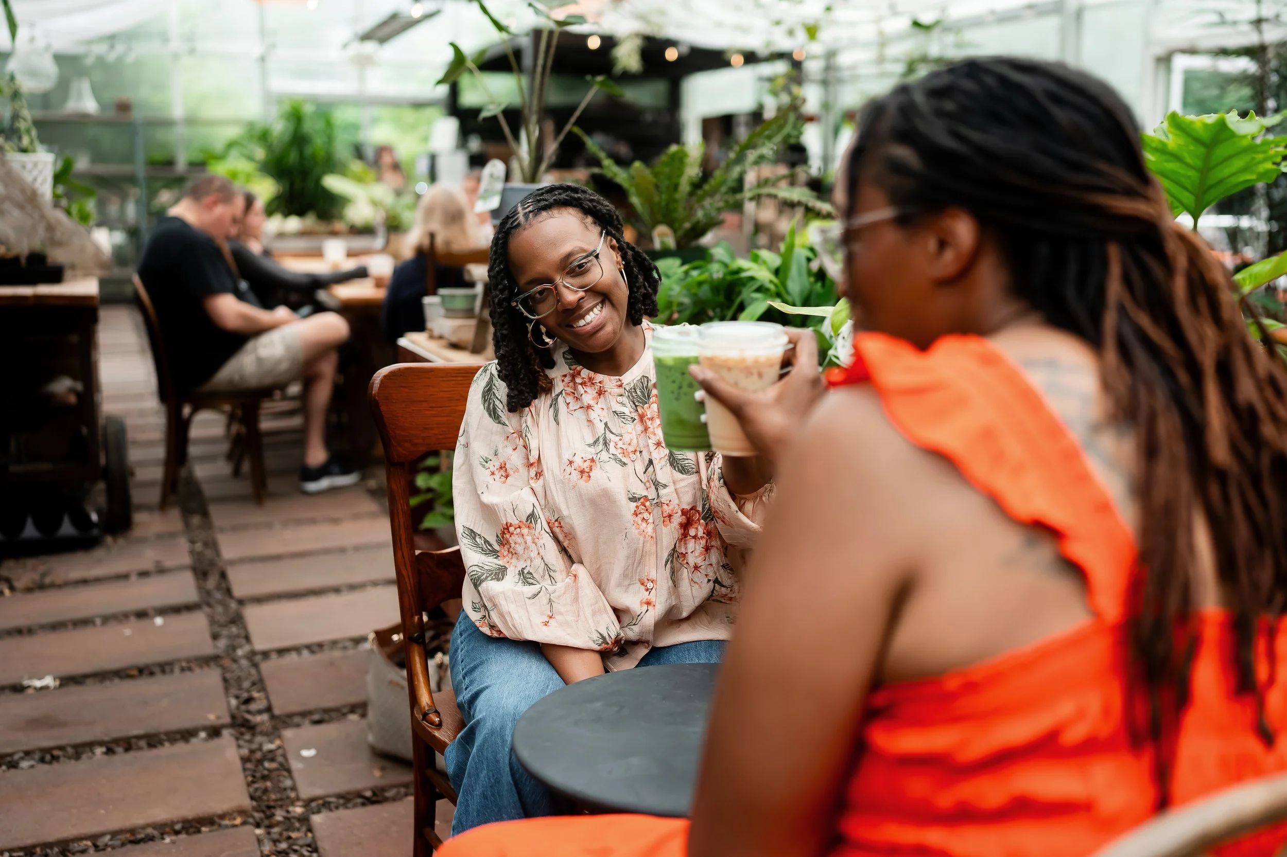 Two women smiling and clinking cups of cold drinks at an indoor or greenhouse setting with lush green plants; blurred background with other people sitting at tables.