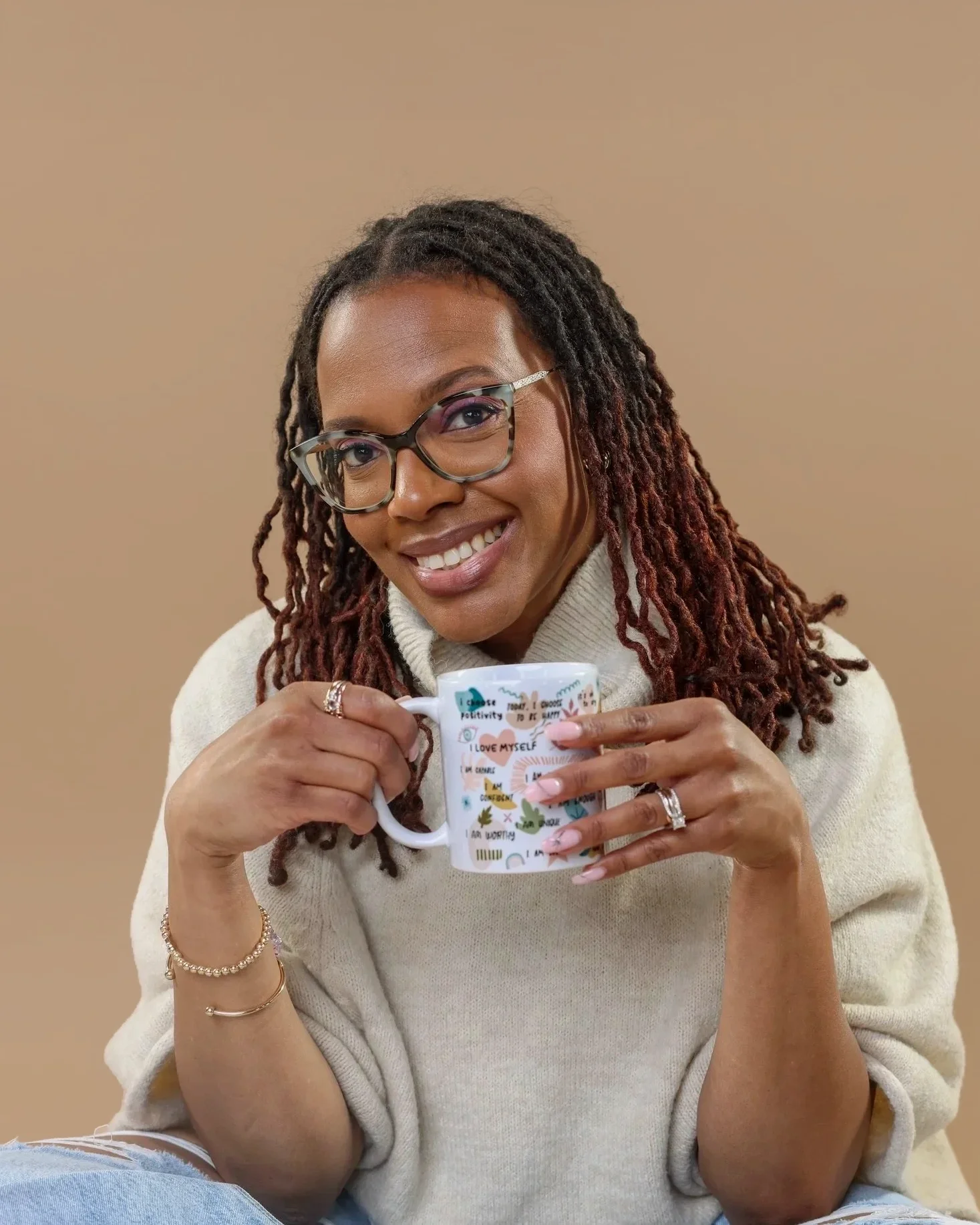 A woman with glasses and dreadlocks holding a colorful coffee mug, smiling, wearing a beige sweater, against a beige background.