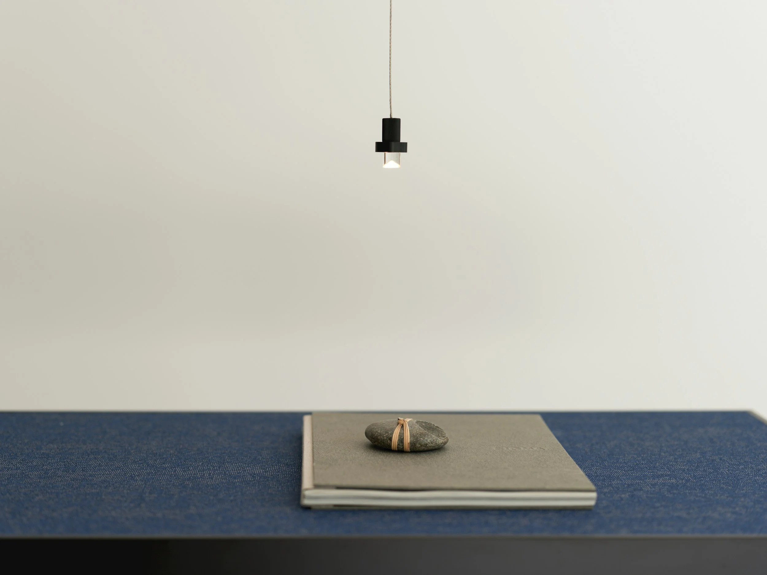 A minimalistic desk with a blue textured surface, a gray notebook, and a decorative stone tied with a beige rubber band. A single hanging light fixture is above the desk.