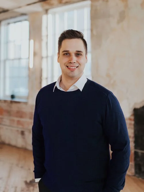 Man in a blue sweater standing in a rustic room with large windows.