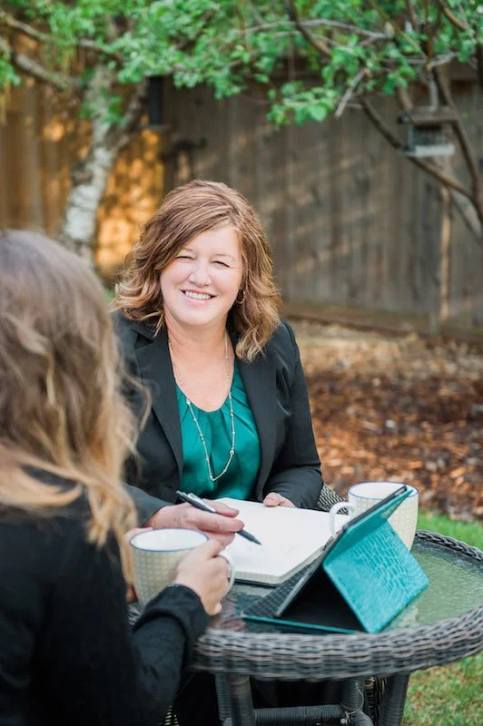 Two women sitting at an outdoor table, one with a notebook and pen, the other holding a cup, with a tablet on the table.
