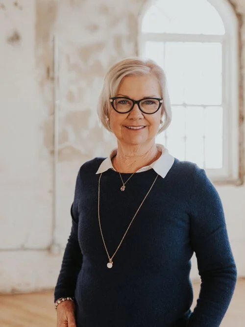 Smiling elderly woman wearing glasses and a navy blue sweater stands in front of a rustic, light-filled background.