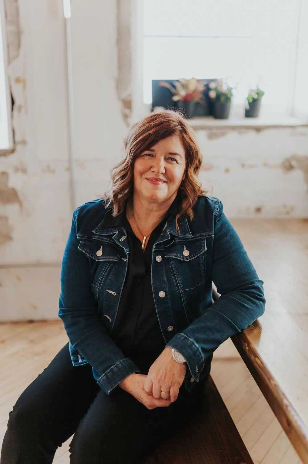 Woman, Lorraine Lawrence, a leadership coach, in denim jacket sitting on a wooden bench indoors.