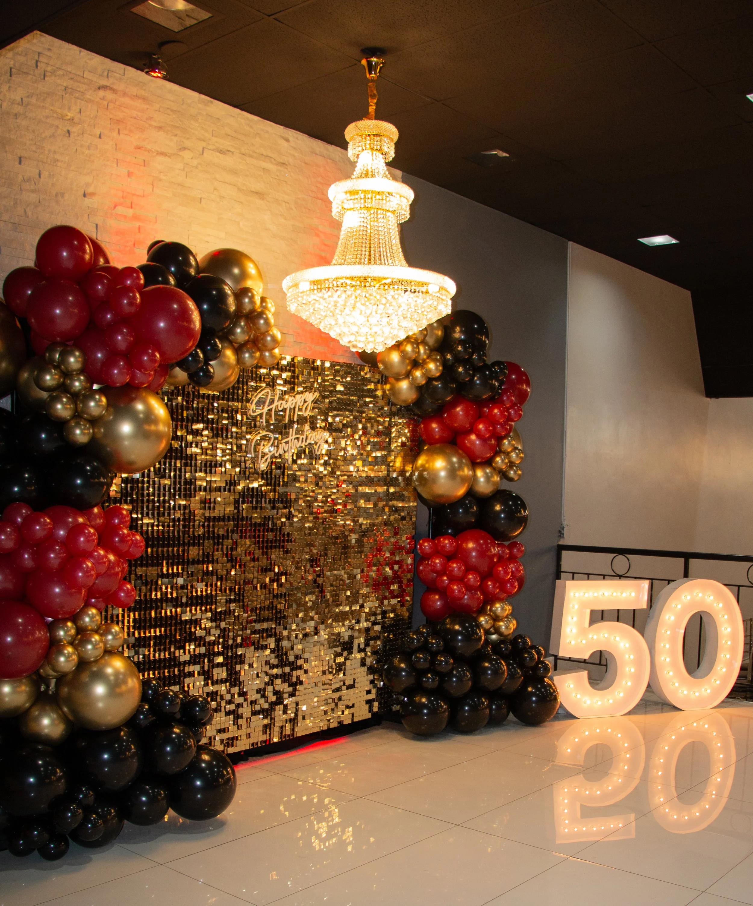 A birthday party scene with a chandelier, a backdrop of gold sequins with "Happy Birthday" sign, clusters of red, black, and gold balloons, and illuminated "50" numbers on a reflective floor.