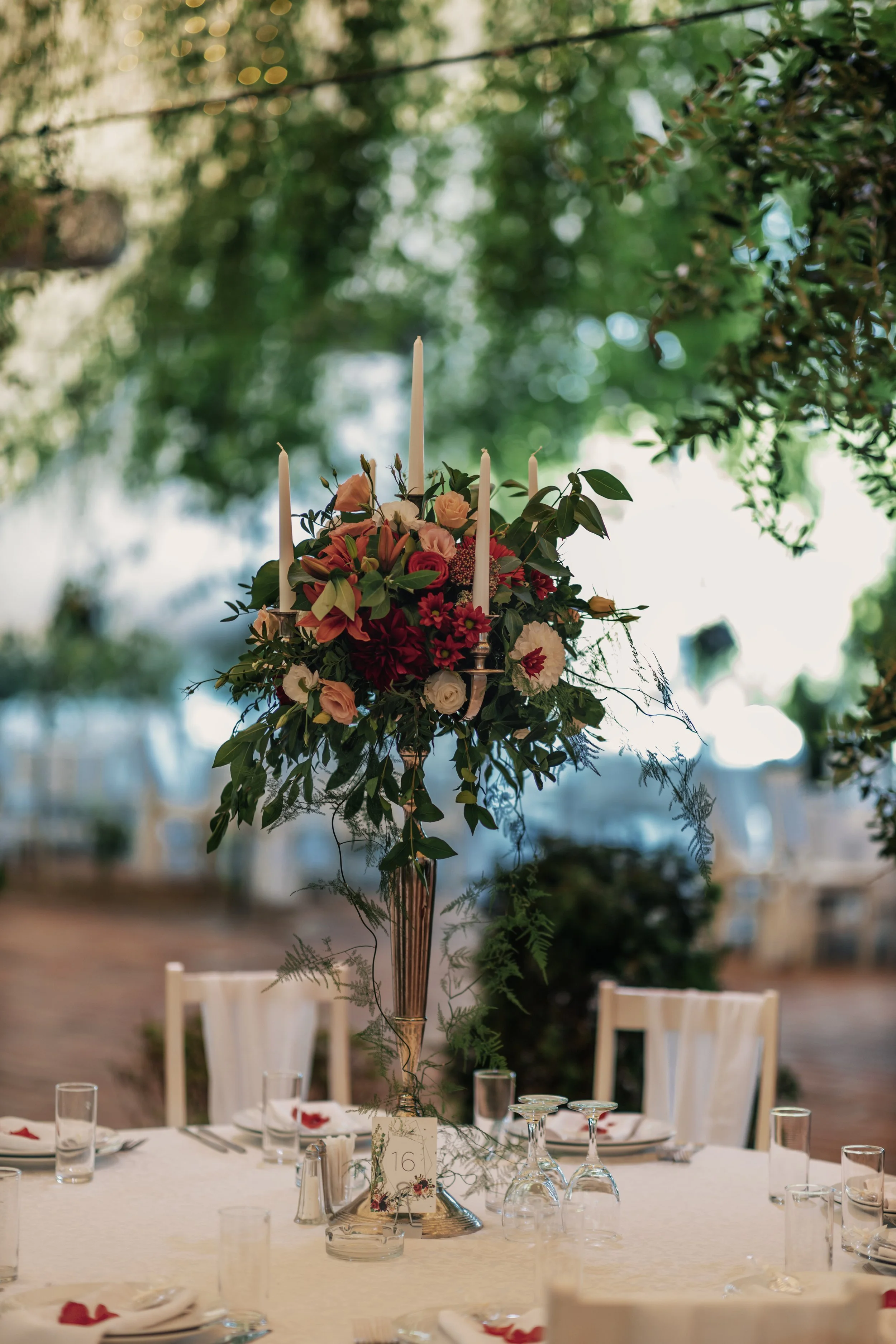 A decorated banquet table with a tall floral centerpiece featuring pink, red, and white flowers and four white candles, set outdoors with chairs and glassware around the table.