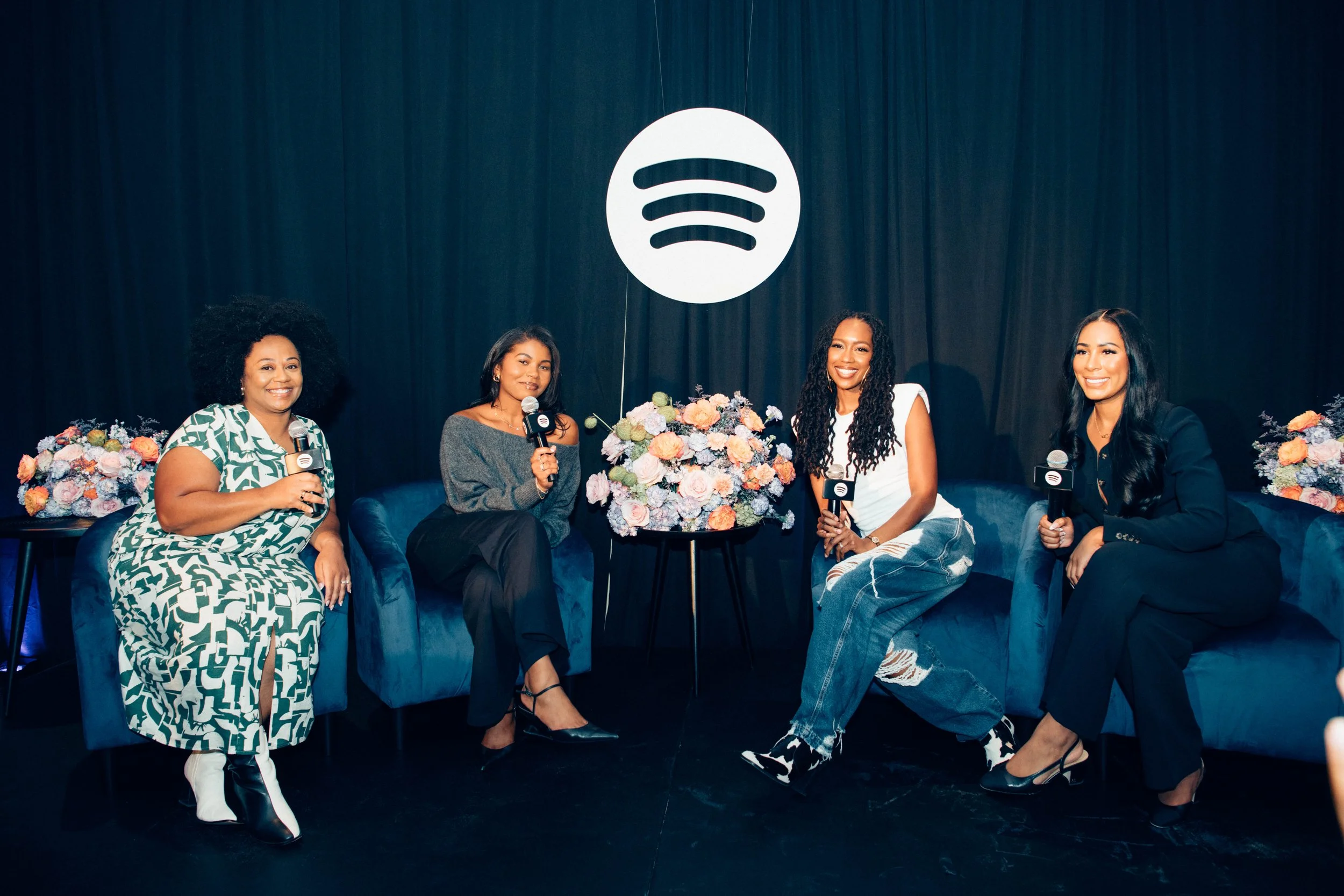 Four women sitting on a blue sofa with microphones, in front of a black curtain with a white Spotify logo hanging above, surrounded by floral arrangements.