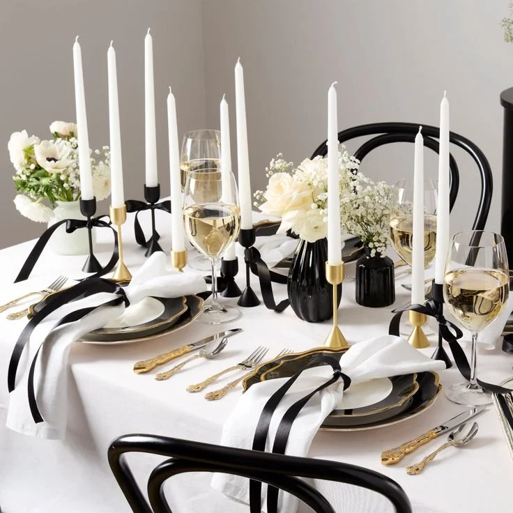 Elegant dinner table setting with black and gold accents, white tablecloth, floral centerpieces, cream and champagne glasses, and tall white candles.