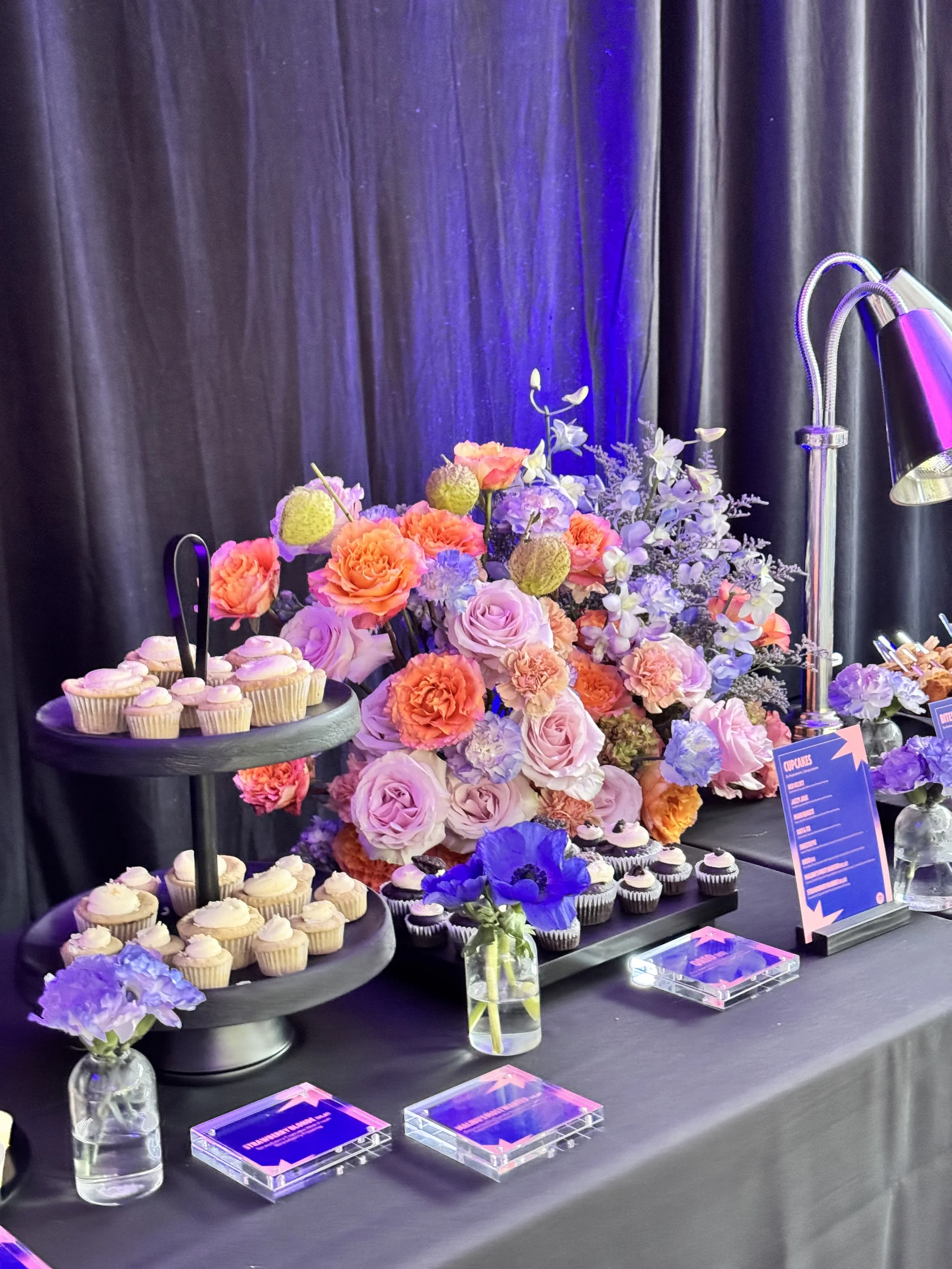 A dessert table decorated with a large arrangement of pink, purple, and orange flowers, cupcakes on a black tiered stand, and small flowers in glass vases. Purple lighting accents the arrangement against a black curtain backdrop.