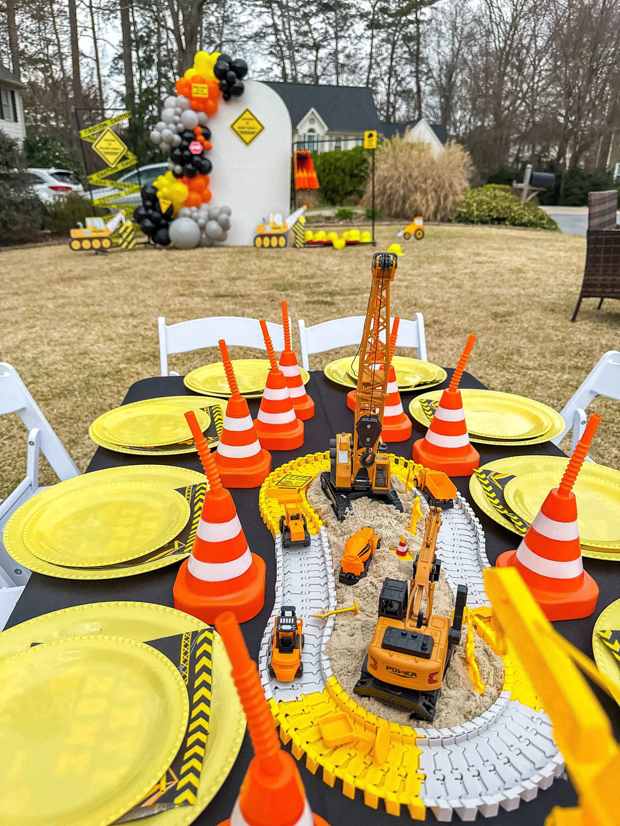 Construction-themed party table with toy construction vehicles, orange traffic cones, and yellow plates, with a construction zone backdrop decorated with balloons and caution tape.