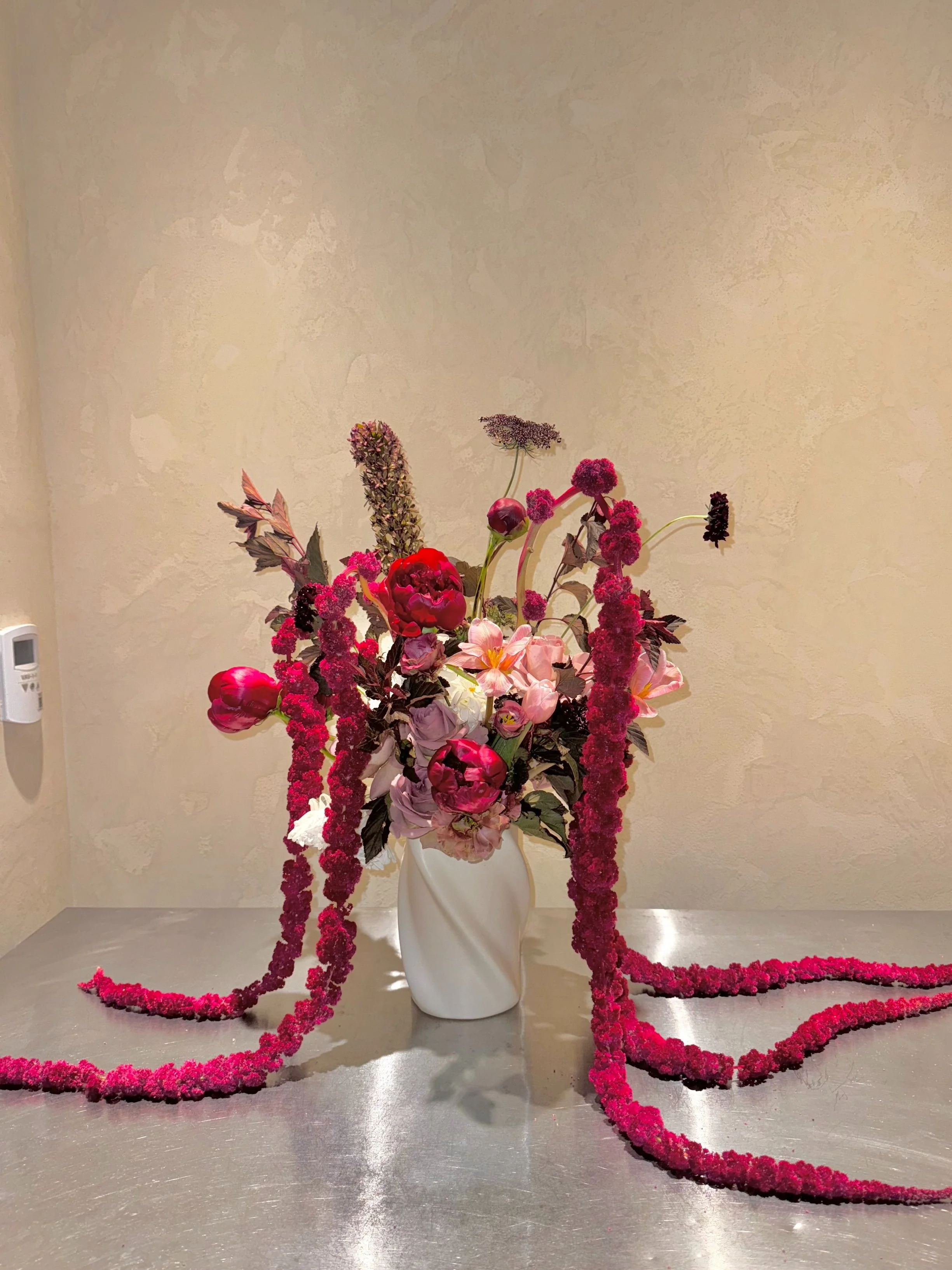 A floral arrangement in a white vase featuring pink and deep red flowers, with some long, trailing pink flower garlands draped over a metallic surface, set against a beige wall.