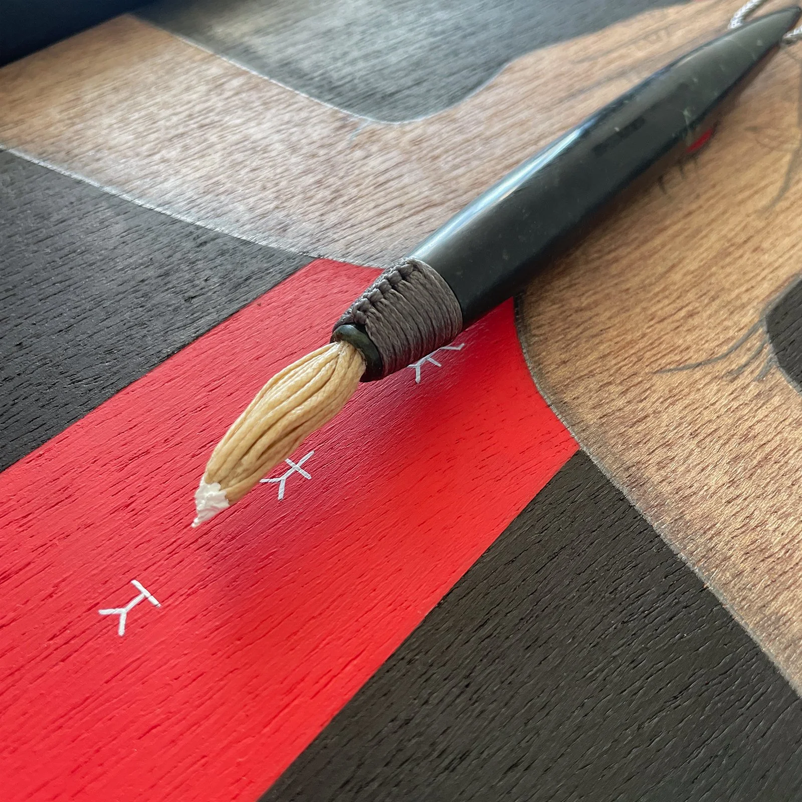 Close-up of the painted bristles on Pounamu carved paintbrush in front of a painting with a red stripe, black background and white symbols.