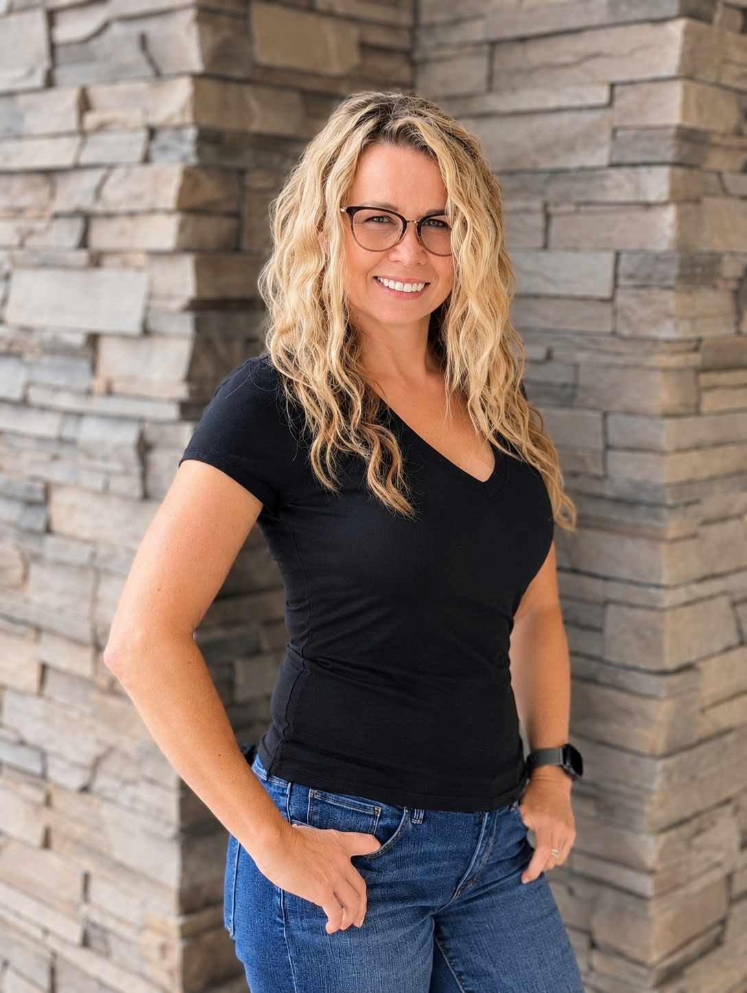 A woman with blonde, curly hair and glasses, wearing a black t-shirt and jeans, standing against a stone wall.