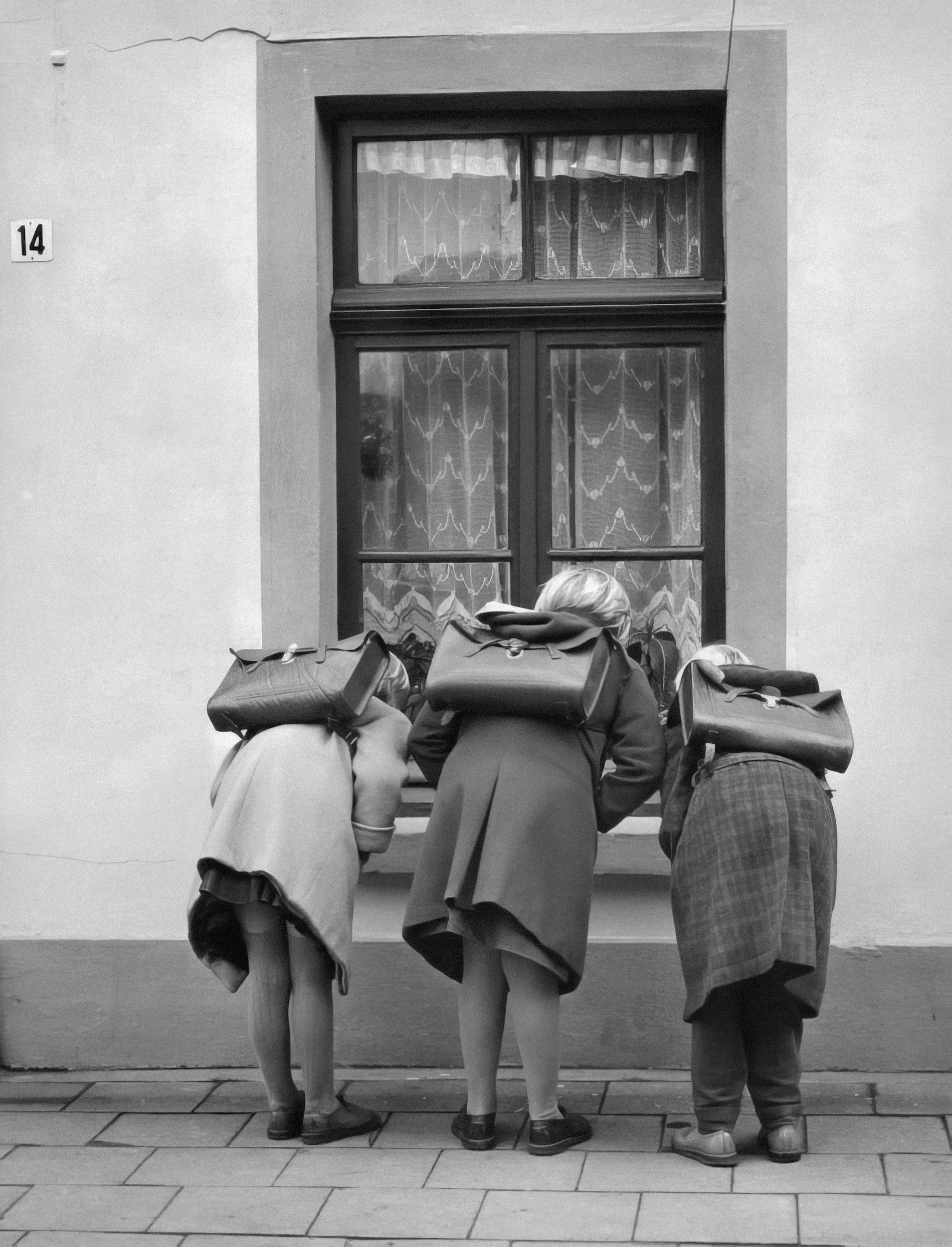 Three women with backpacks looking into a house window, seen from the back, in black and white.