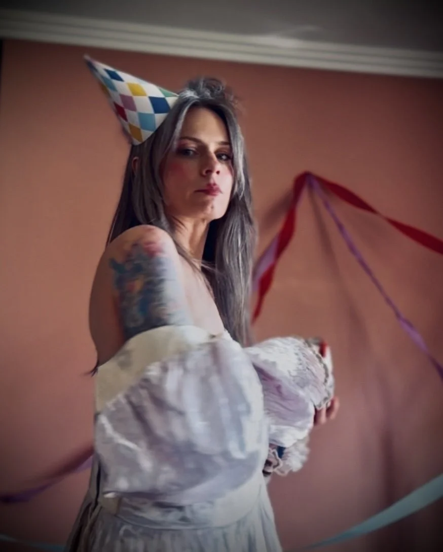 Woman with gray hair wearing a colorful birthday hat at a celebration, holding a gift, with streamers and balloons in the background.