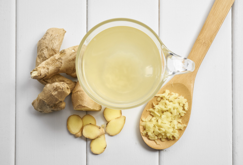 Fresh ginger root, ginger slices, grated ginger in wooden spoon, and cup of ginger tea on white wooden surface.