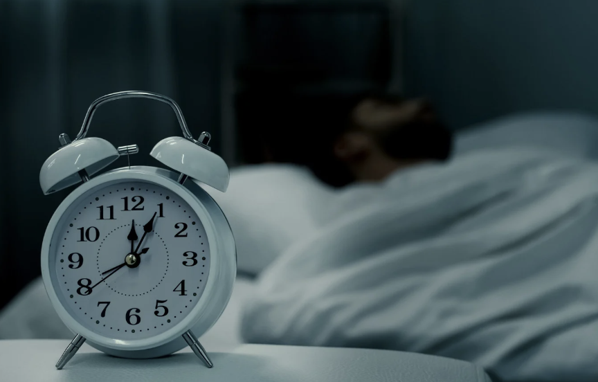 Close-up of a white alarm clock showing 6:30 with a blurred person sleeping in the background
