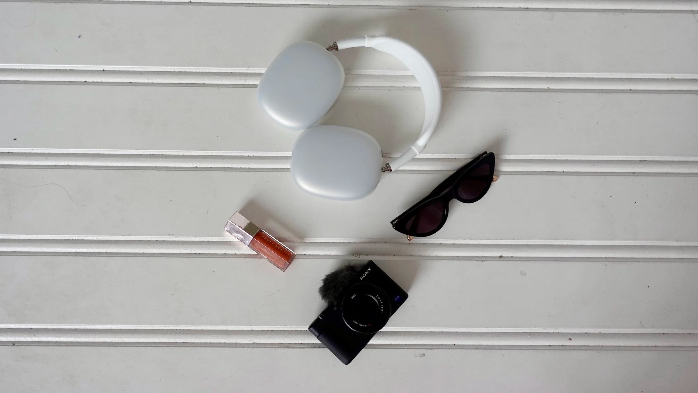 A white surface with horizontal grooves holding a pair of white headphones, black sunglasses, a tube of lip gloss, a compact camera, and a furry microphone windscreen.