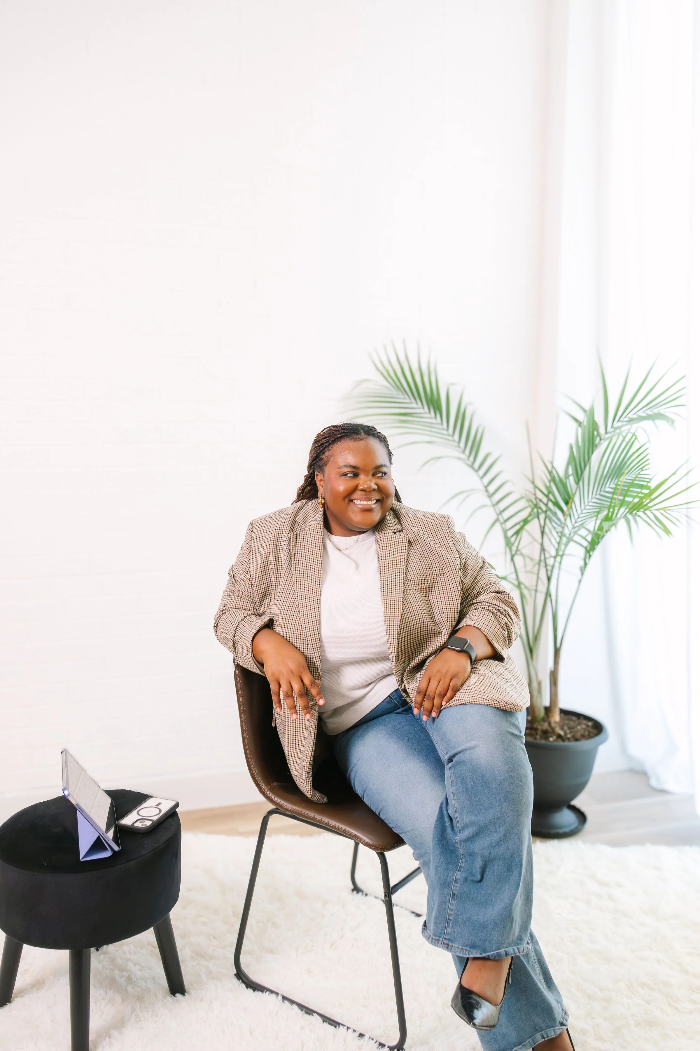 A cheerful woman with dark braided hair, wearing a beige checkered blazer, white t-shirt, and blue jeans, sitting on a brown chair with her legs crossed in a bright room with white walls and a large potted plant behind her.