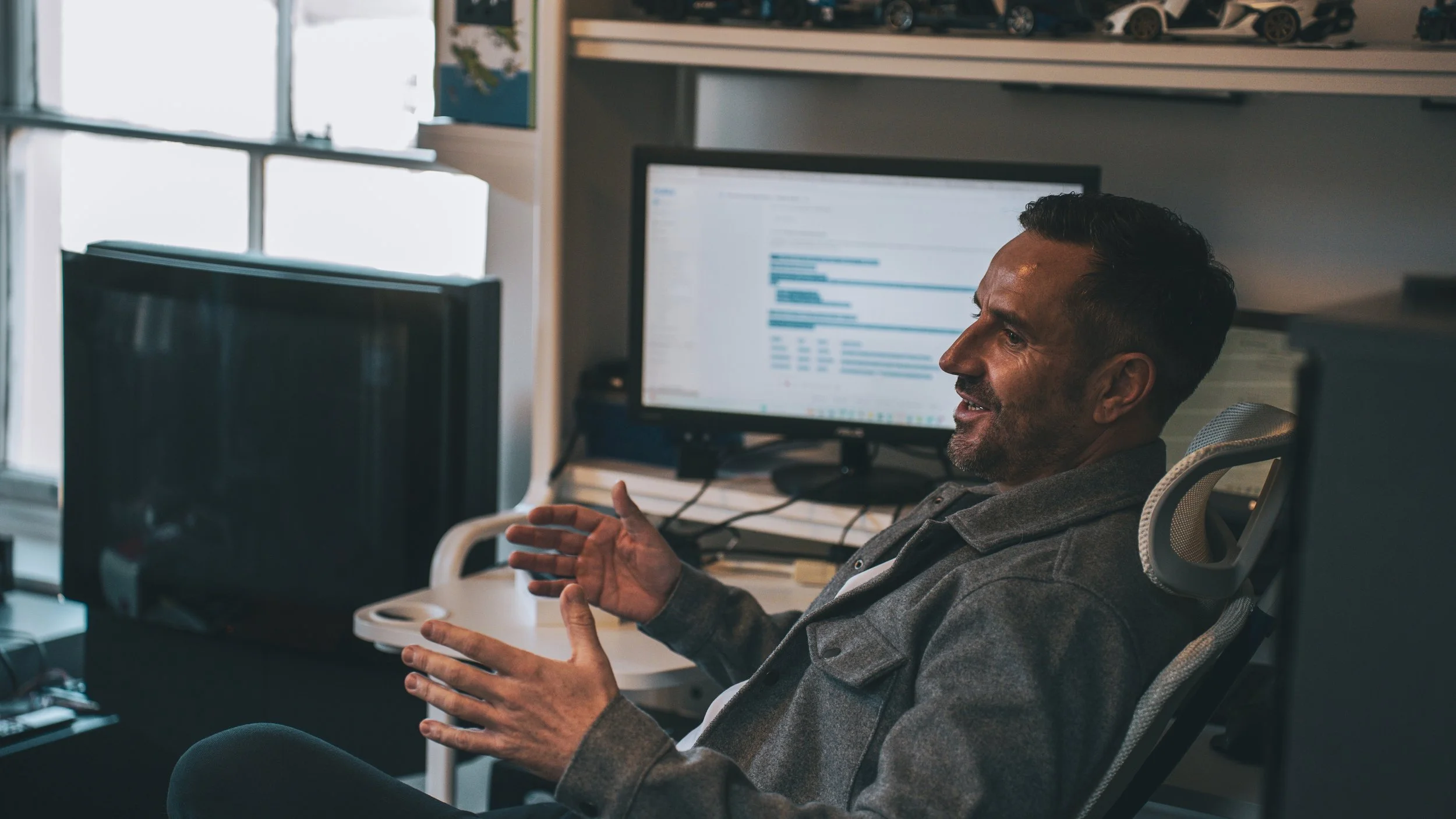 Kenny Skelton, sitting in an office, engaging in conversation while gesturing with his hands. He is near a computer monitor displaying a spreadsheet or email and there are other office equipment around him.