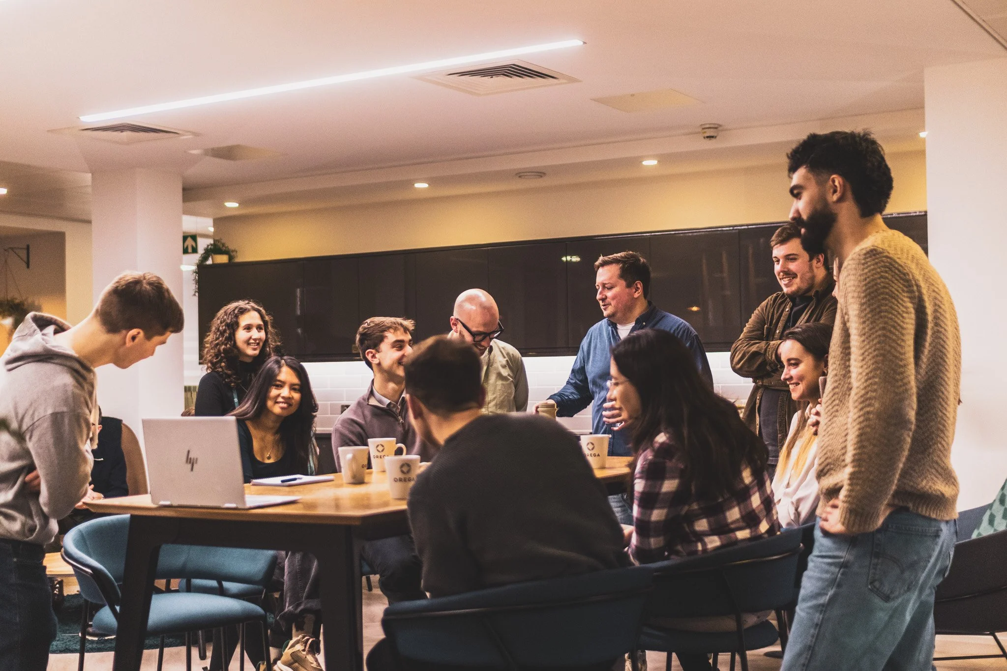 A diverse group of young adults gathered around a table in an office or cafe, laughing and engaging in conversation.