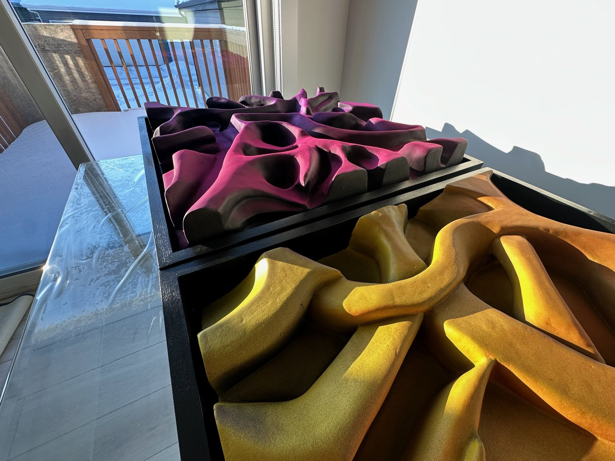 Abstract geometric sculptures in purple and yellow displayed indoors, with a snowy outdoor scene visible through a glass door in the background.