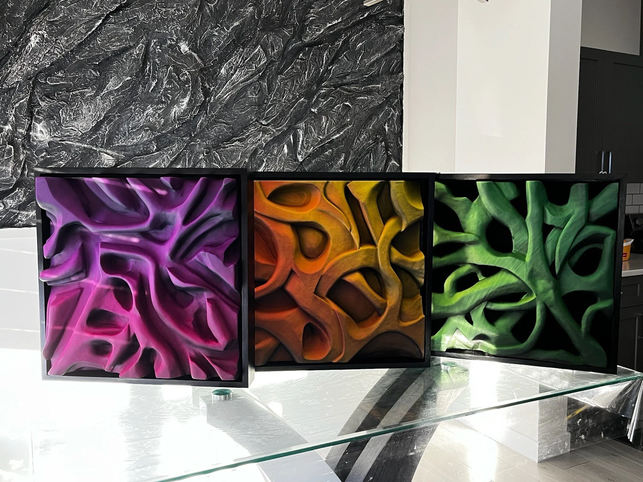 Three abstract 3D artworks in vibrant colors: purple, orange, and green, displayed on a glass surface against a textured wall.