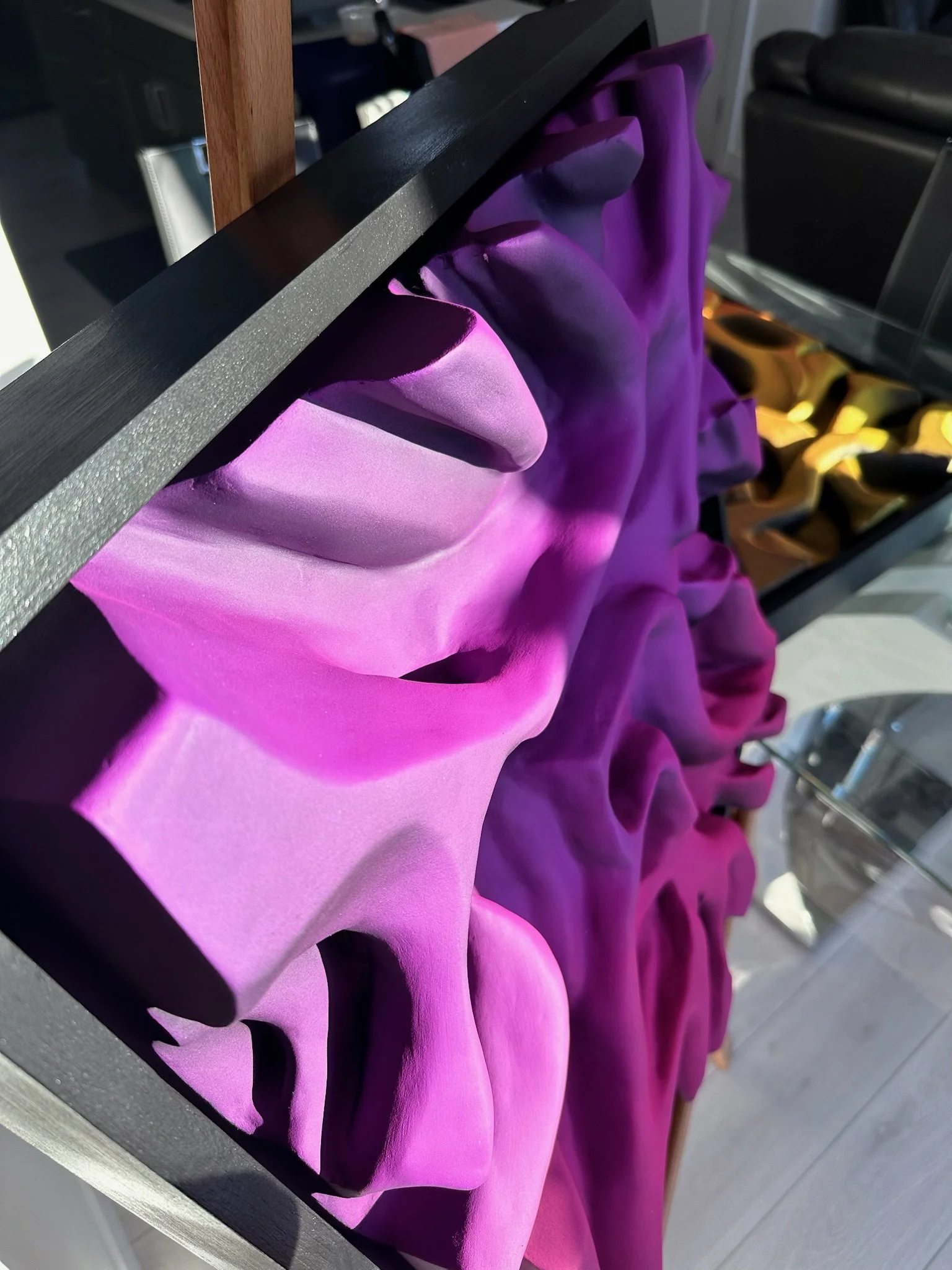 Close-up of a purple and pink textured sculpture with undulating shapes and a black frame, placed on a surface with sunlight casting shadows.