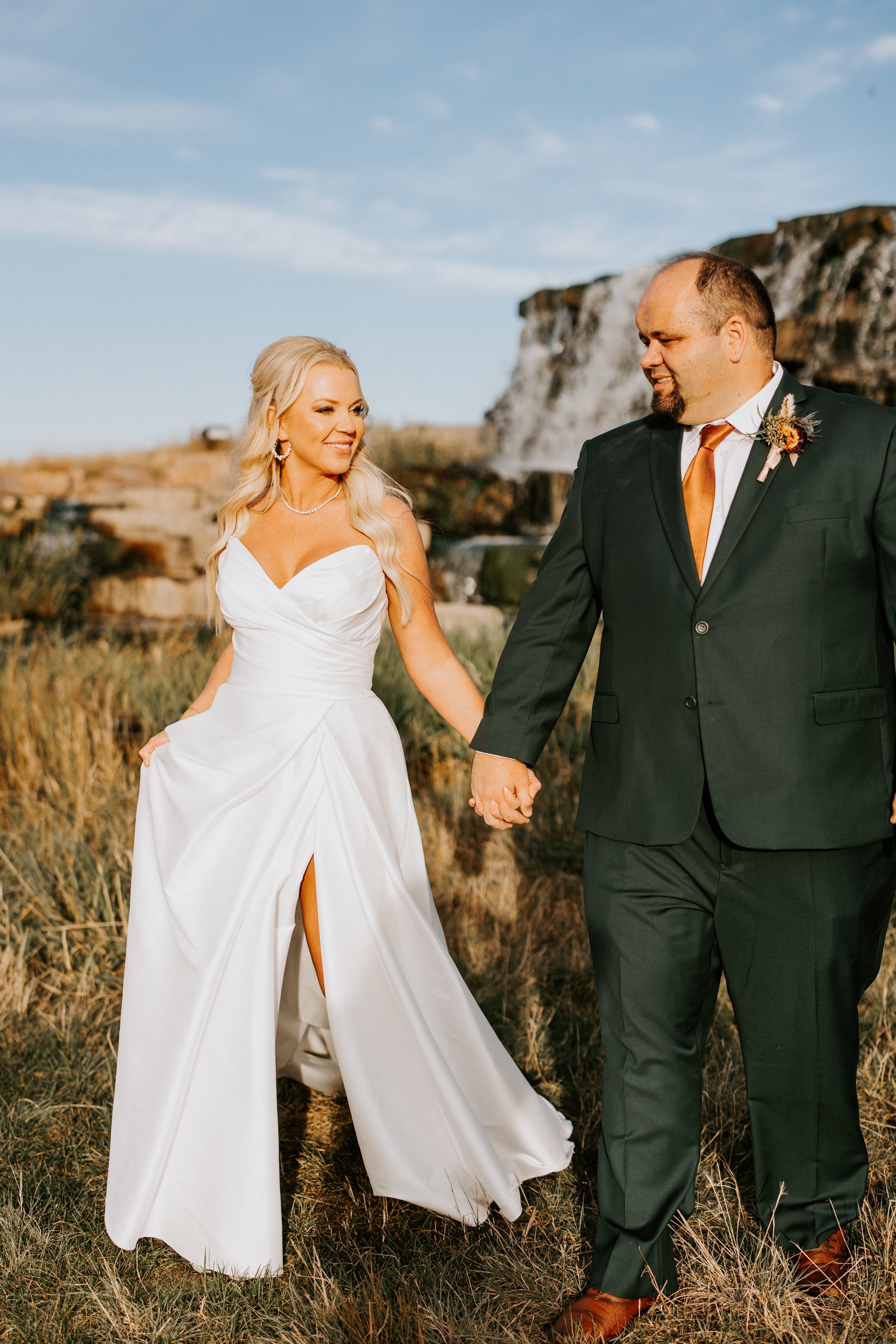 A bride and groom holding hands and walking outdoors in a field with rocks and grass, under a partly cloudy sky, during their wedding.