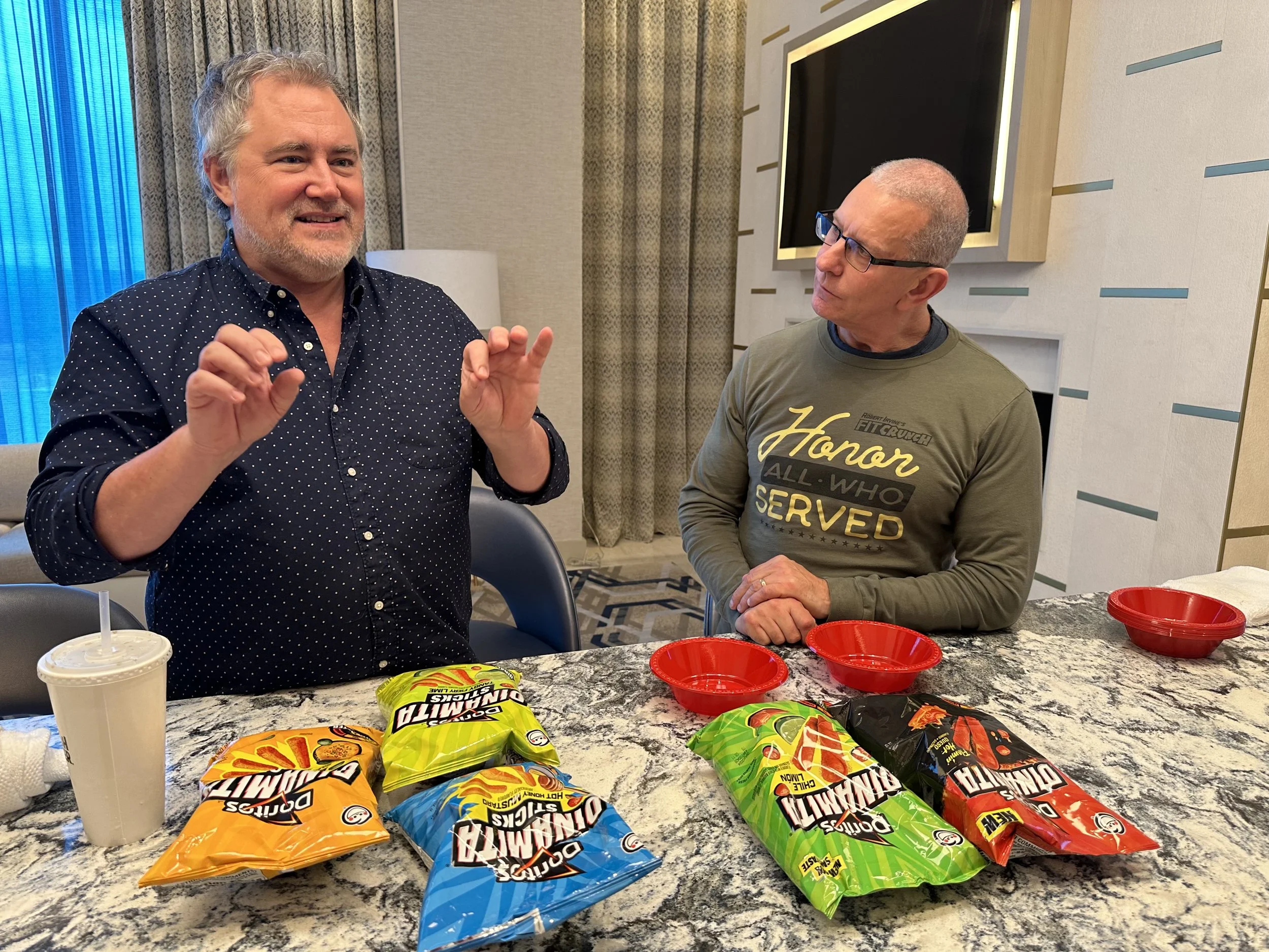 Two men standing behind a marble table with snack bags and red bowls, engaging in conversation. One man has gray hair and is wearing a navy blue polka-dot shirt; the other man is bald, wearing glasses, and a green shirt that reads 'Honor all who served.'