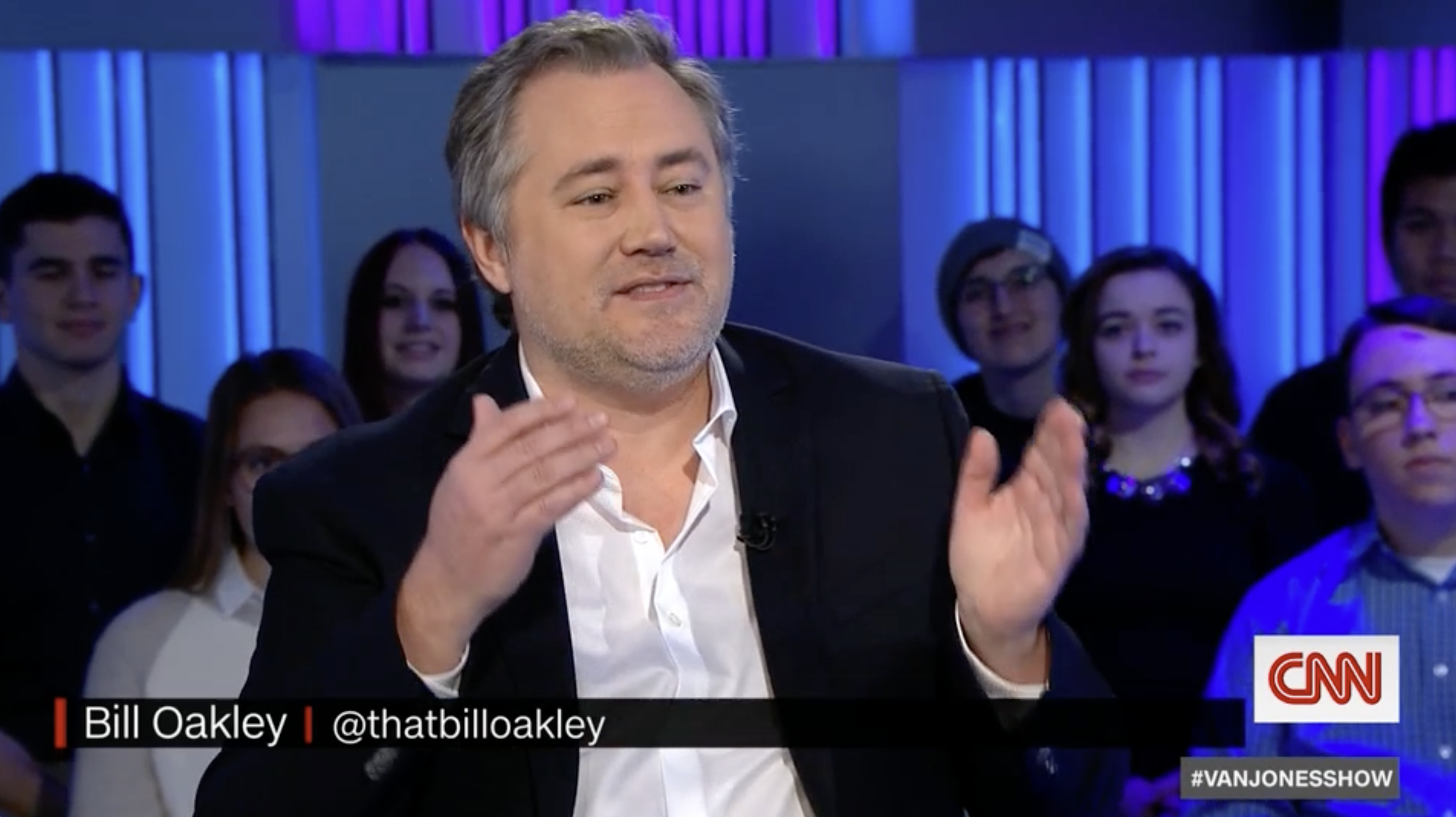 Bill Oakley speaking during an interview on CNN with an audience behind him, featuring young adults and blue and purple lighting.