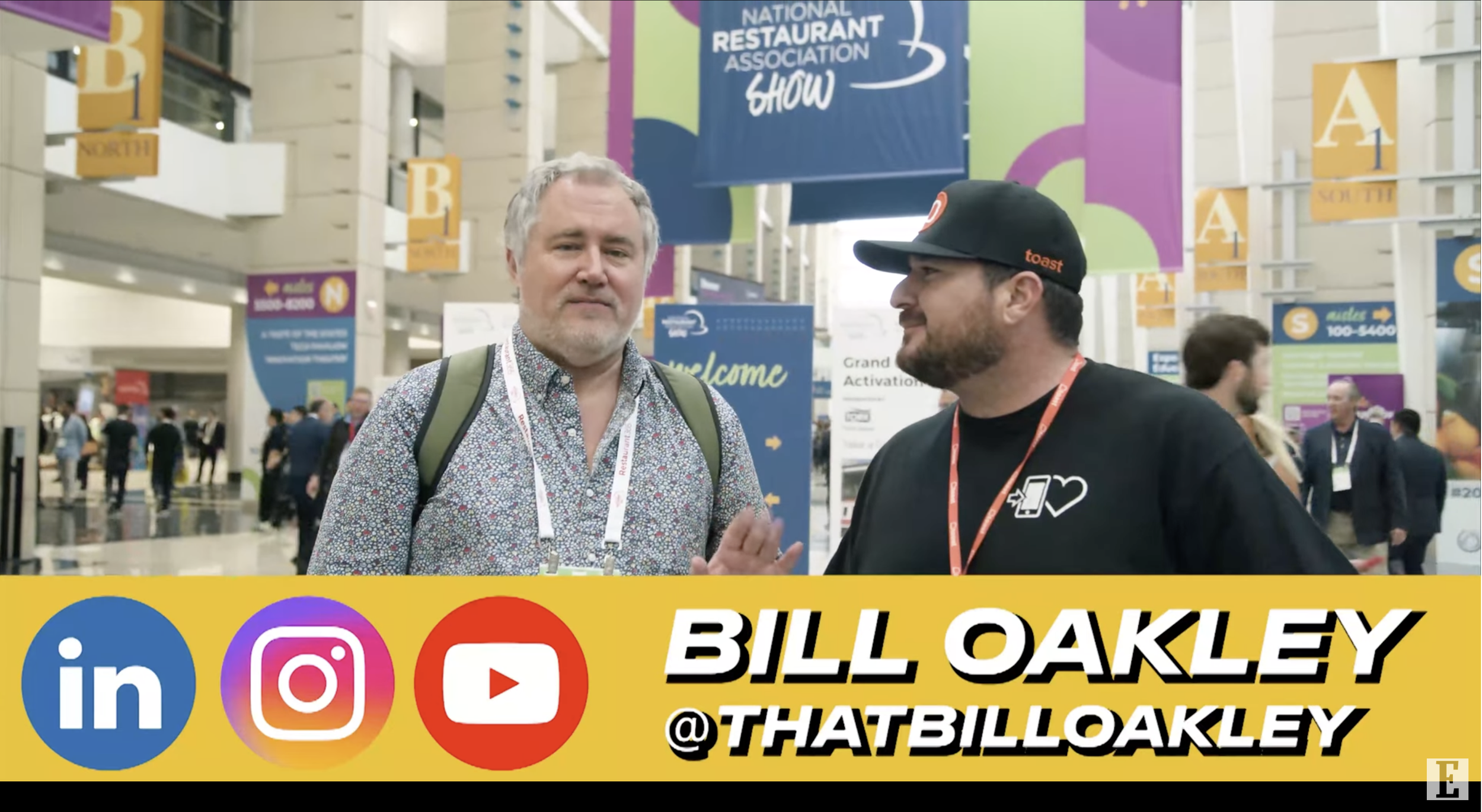 A man with a gray beard and a floral shirt standing next to a man with a beard, wearing a black hat and T-shirt, at a busy convention center with banners and signs for a restaurant association show in the background.