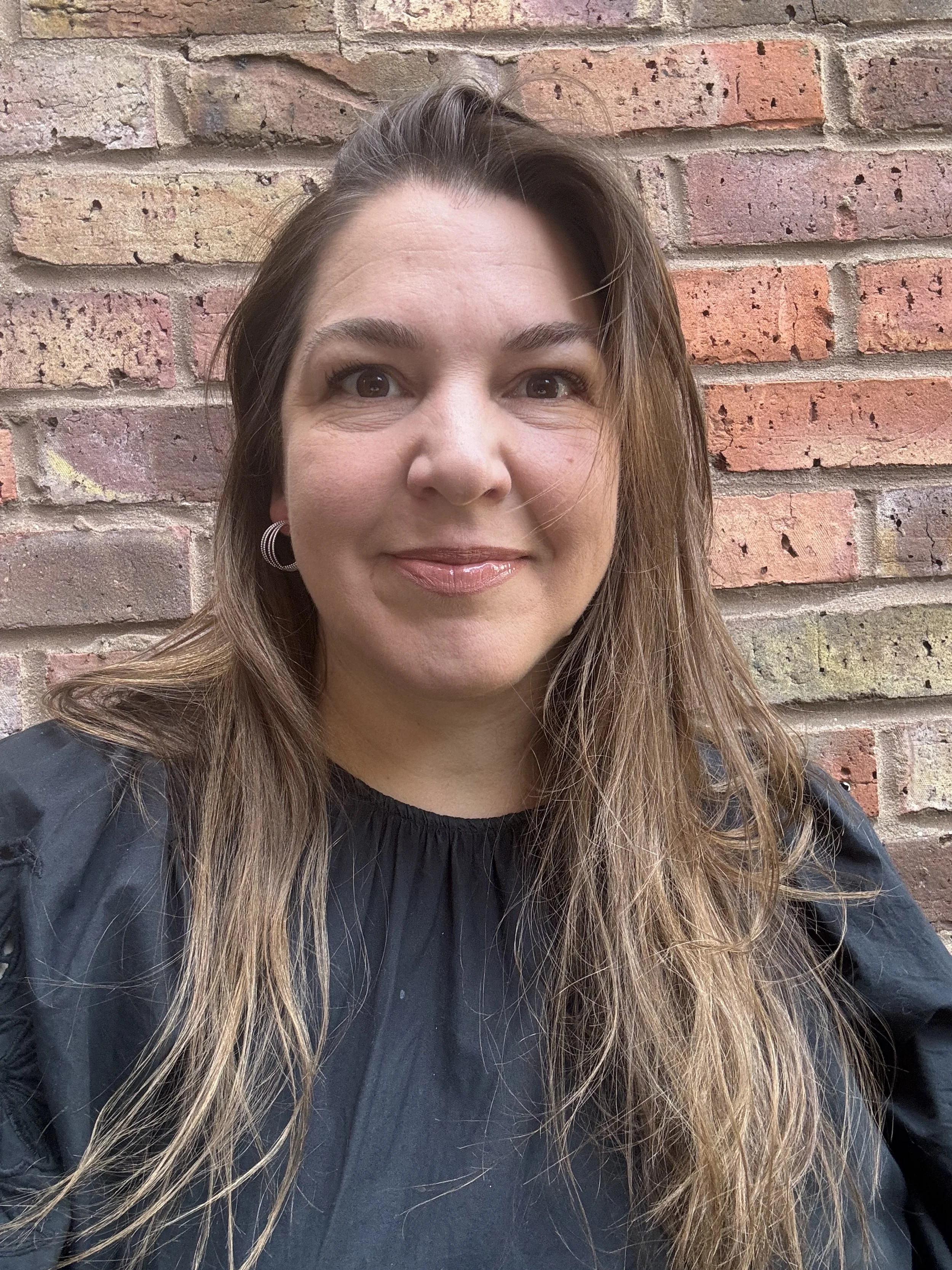 A woman with long brown hair and hoop earrings taking a selfie in front of a brick wall.