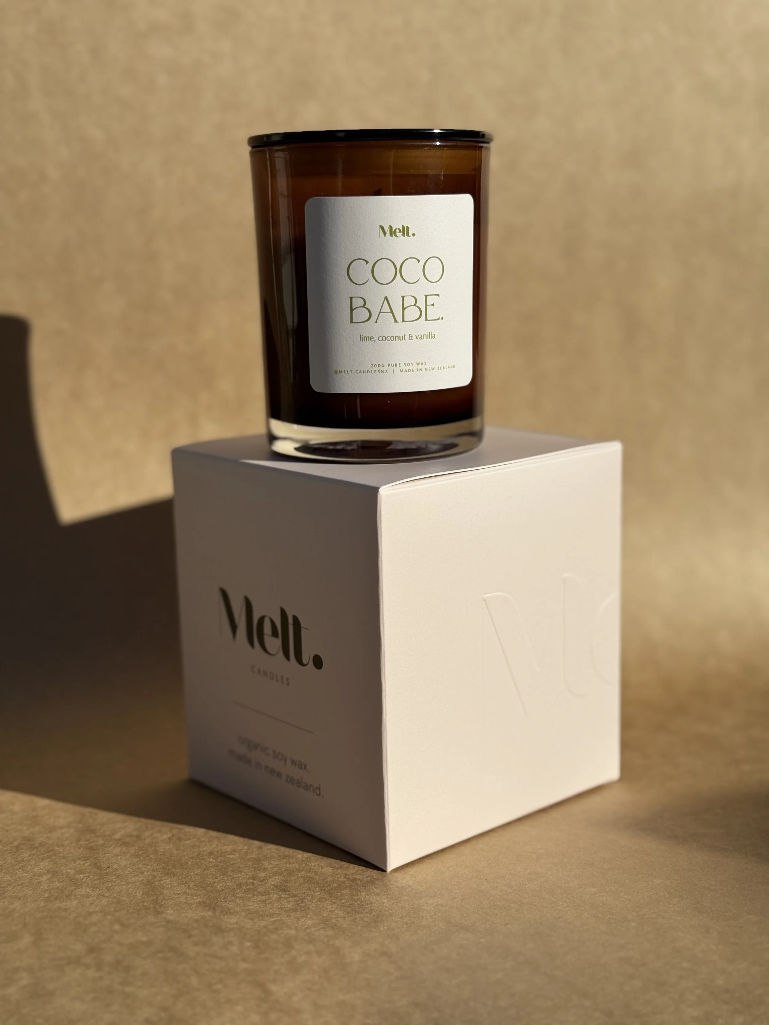 A brown glass candle jar labeled 'Coco Babe' with lime, coconut, and vanilla scent, placed on a white box with embossed 'Velt' branding, on a brown background.