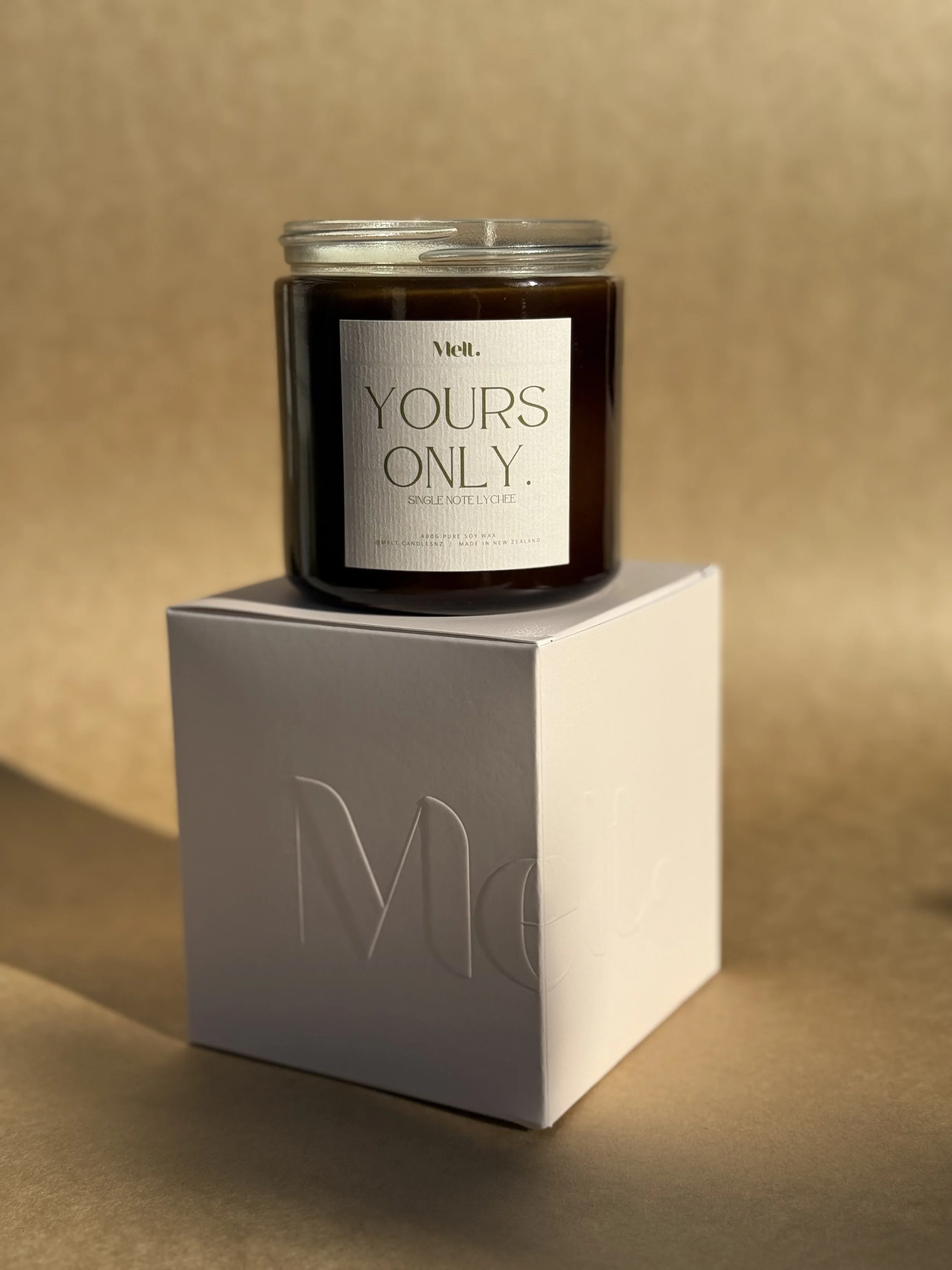 A black candle in a glass jar with a white label that reads 'YOURS ONLY.' and 'Melt.' is placed on top of a white box with embossed text.