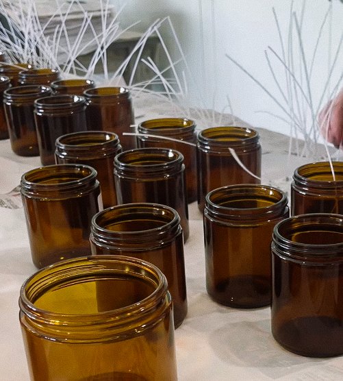 Multiple amber glass jars on a table, some with wax sticks inside, likely for making candles.