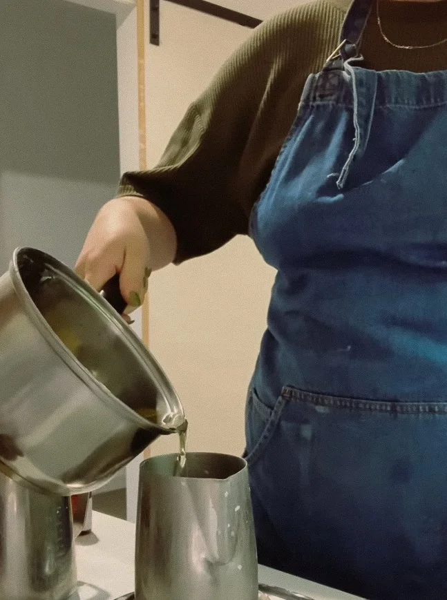 Person pouring liquid from a metal pot into a metal cup, wearing a green shirt and blue apron.