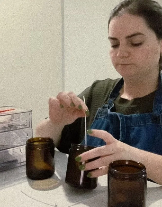 A woman with dark hair and painted nails is using a dropper to transfer liquid into a small black bottle. She is sitting at a table with two brown glass jars and some transparent storage containers nearby.