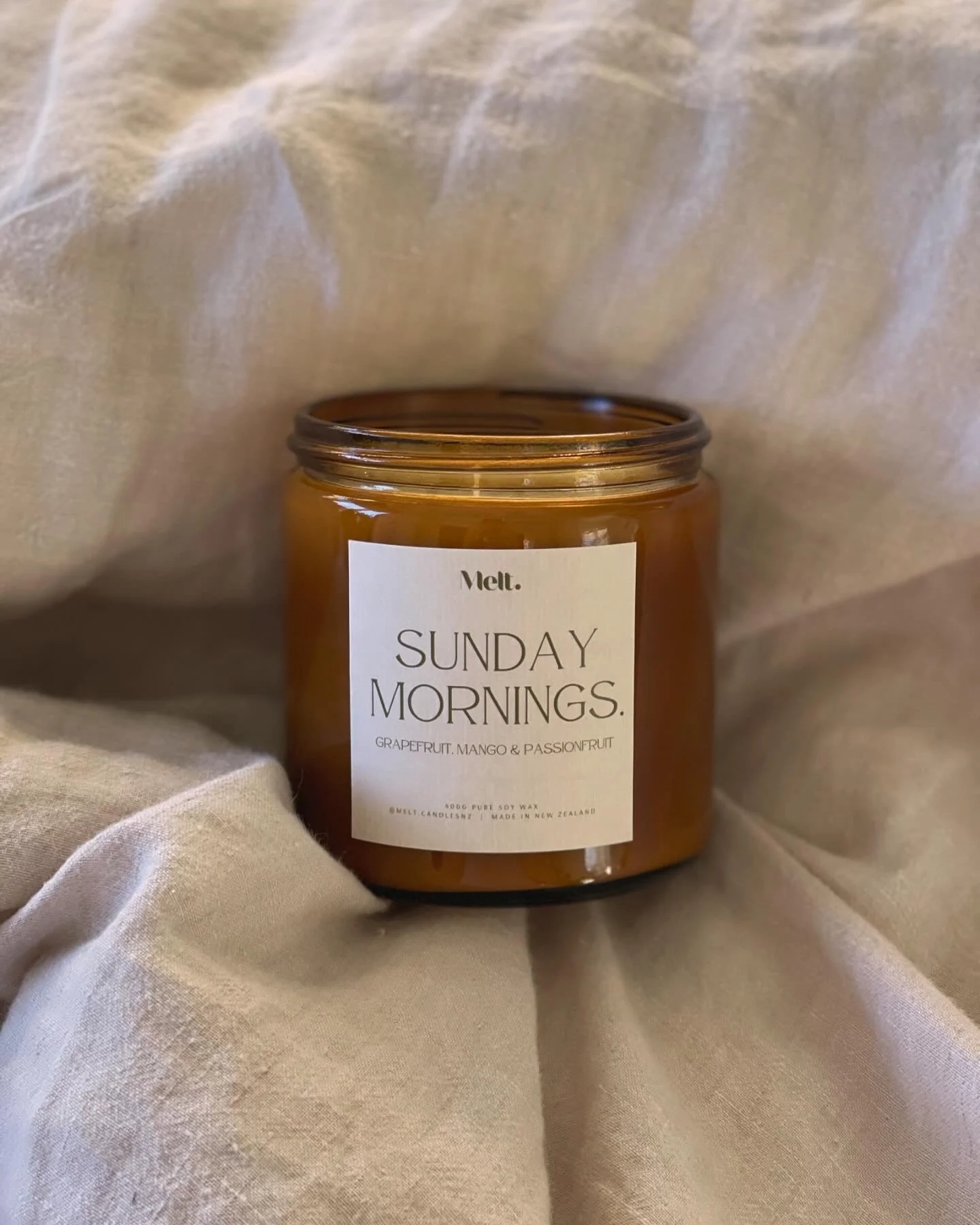 A candle jar labeled 'Sunday Mornings' with grapefruit, mango, and passionfruit scent on a soft beige fabric background.