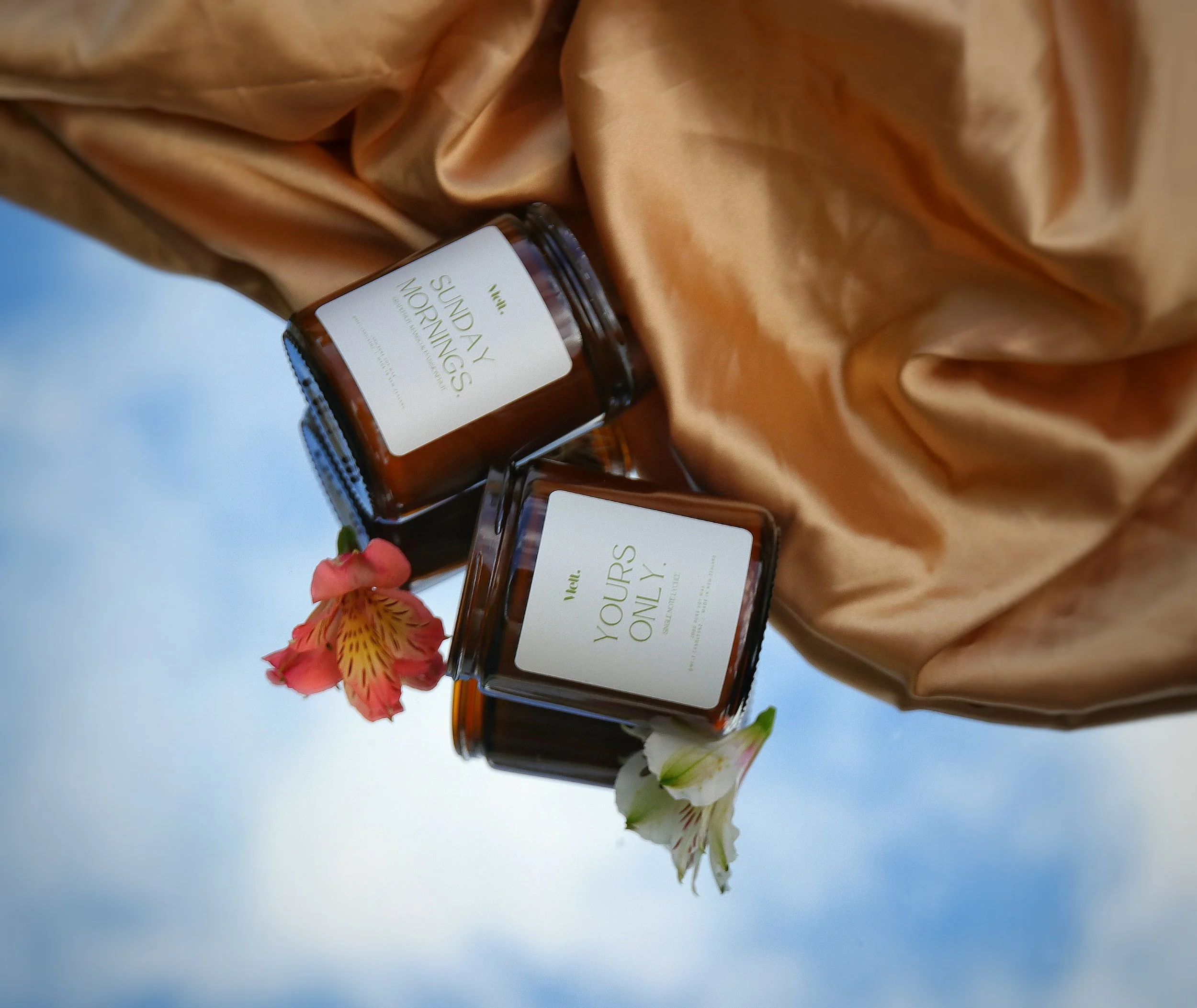 Two amber-colored candles with white labels, one labeled 'Your Yoours Only' and the other 'Sunday Mornings,' adorned with pink and white flowers, set on a beige satin fabric with a blue sky background.