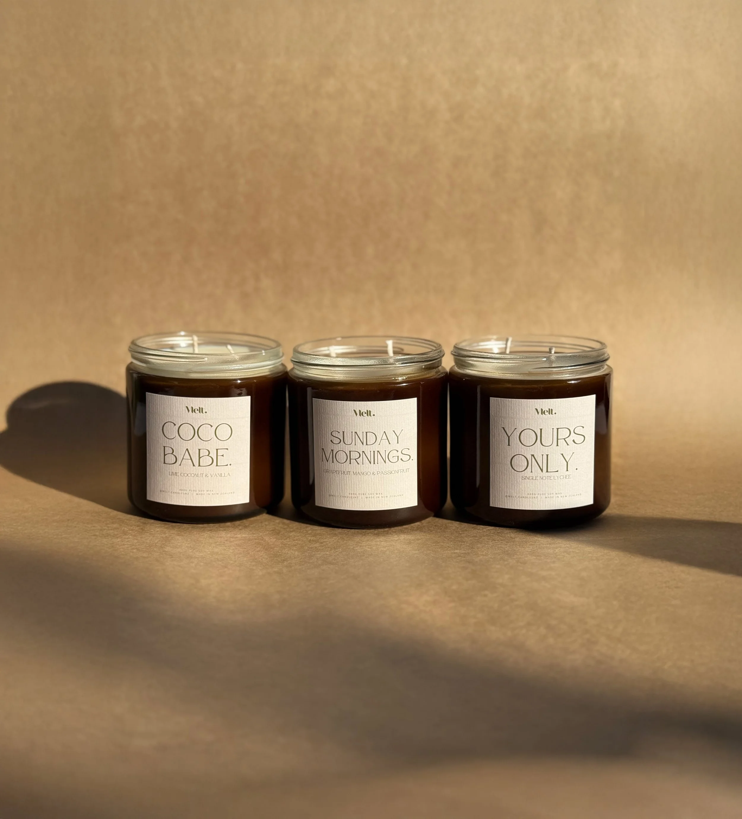 Three candles in glass jars with minimalist labels, each with a different scent name, placed on a beige surface with soft lighting casting shadows.